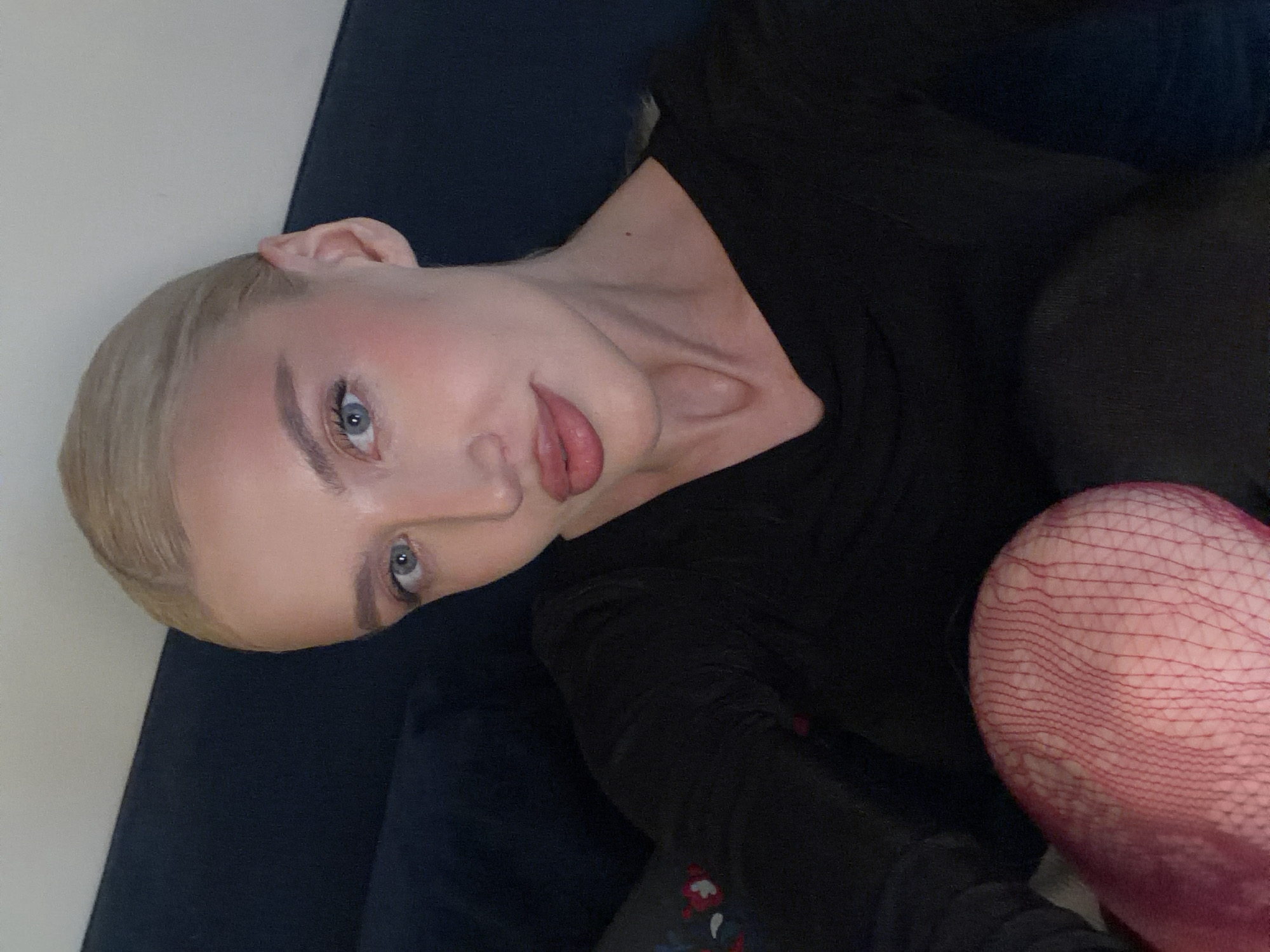 A light-skinned person with blonde hair and blue eyes is lying on a dark blue surface, possibly a couch. They are wearing a black long-sleeved shirt and red fishnet stockings are visible on their leg, which is bent toward their chest. The setting appears to be indoors, with a plain white wall in the background. The image does not contain information to confirm a location in Belgrade, Serbia.