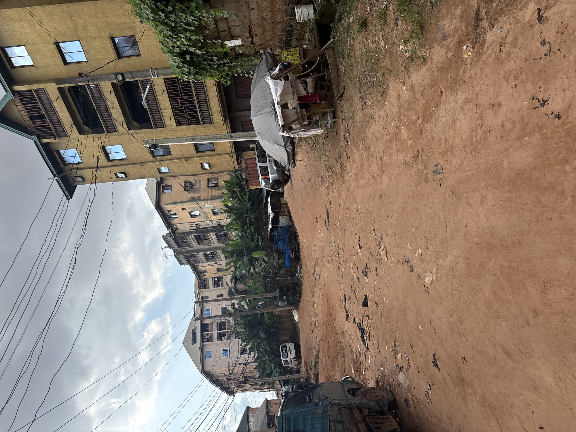 This is a ground-level, eye-level, full shot of a dirt road in a densely populated area of Onitsha, Nigeria. The road is unpaved and appears to be made of reddish-brown soil, littered with various types of debris including plastic bags, paper, and other refuse. On the right side of the road, there is a small roadside stall covered by a grey tarp, propped up by wooden poles. Behind the stall, a concrete wall is visible, partially covered by green vines.
Several multi-story buildings are situated along the left and background of the road, suggesting a residential or mixed-use urban environment. These buildings are of varying heights and architectural styles, many with balconies and visible windows. One prominent building on the right is painted a mustard yellow, with multiple floors and balconies. A silver SUV is parked on the left side of the road, near the yellow building.
In the foreground on the left, the side of a blue utility truck is visible. Further down the road, a white van is parked. A blue flatbed truck is also visible further in the distance. The sky is overcast with a mix of grey clouds and patches of lighter sky. The overall impression is one of a busy, somewhat unkempt urban environment typical of parts of Onitsha.