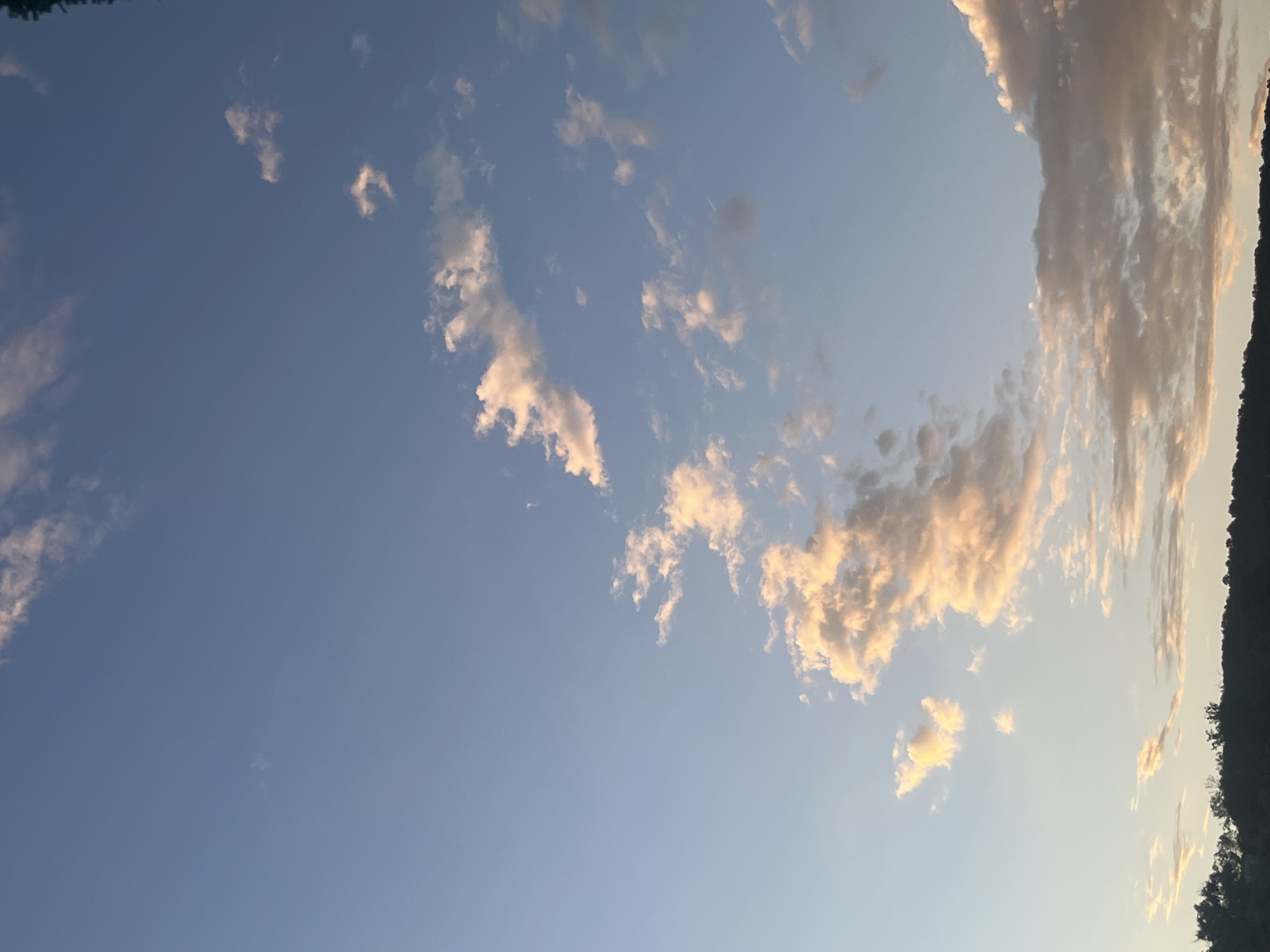 The image depicts a clear sky with scattered clouds, illuminated by the warm hues of what appears to be a sunset or sunrise. The clouds range in color from bright white to soft pink and orange, indicating the angle of the sun. The lower portion of the image shows a dark silhouette of trees and a gentle slope, suggesting a landscape or elevated terrain. The sky itself is a gradient of blue, transitioning from a lighter shade at the horizon to a deeper tone higher up. There are no discernible people, buildings, or specific landmarks that would place the scene within Varna, Bulgaria, or any other particular city. The environment is natural, characterized by sky and landforms.