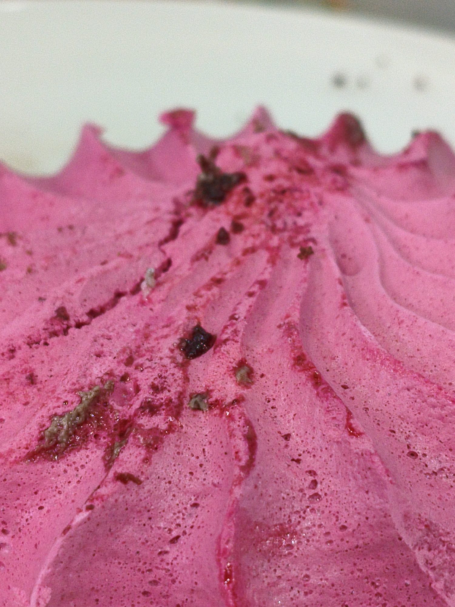 This is a close-up, macro photograph of a pink, whipped confection, possibly frosting or mousse, with visible textured ridges and small dark specks, likely bits of fruit or chocolate. The background is out of focus and appears to be a white surface. There is no indication of Orenburg, Russia, or any specific location within a city. The image focuses solely on the texture and color of the food item. There are no people, actions, or interactions depicted.