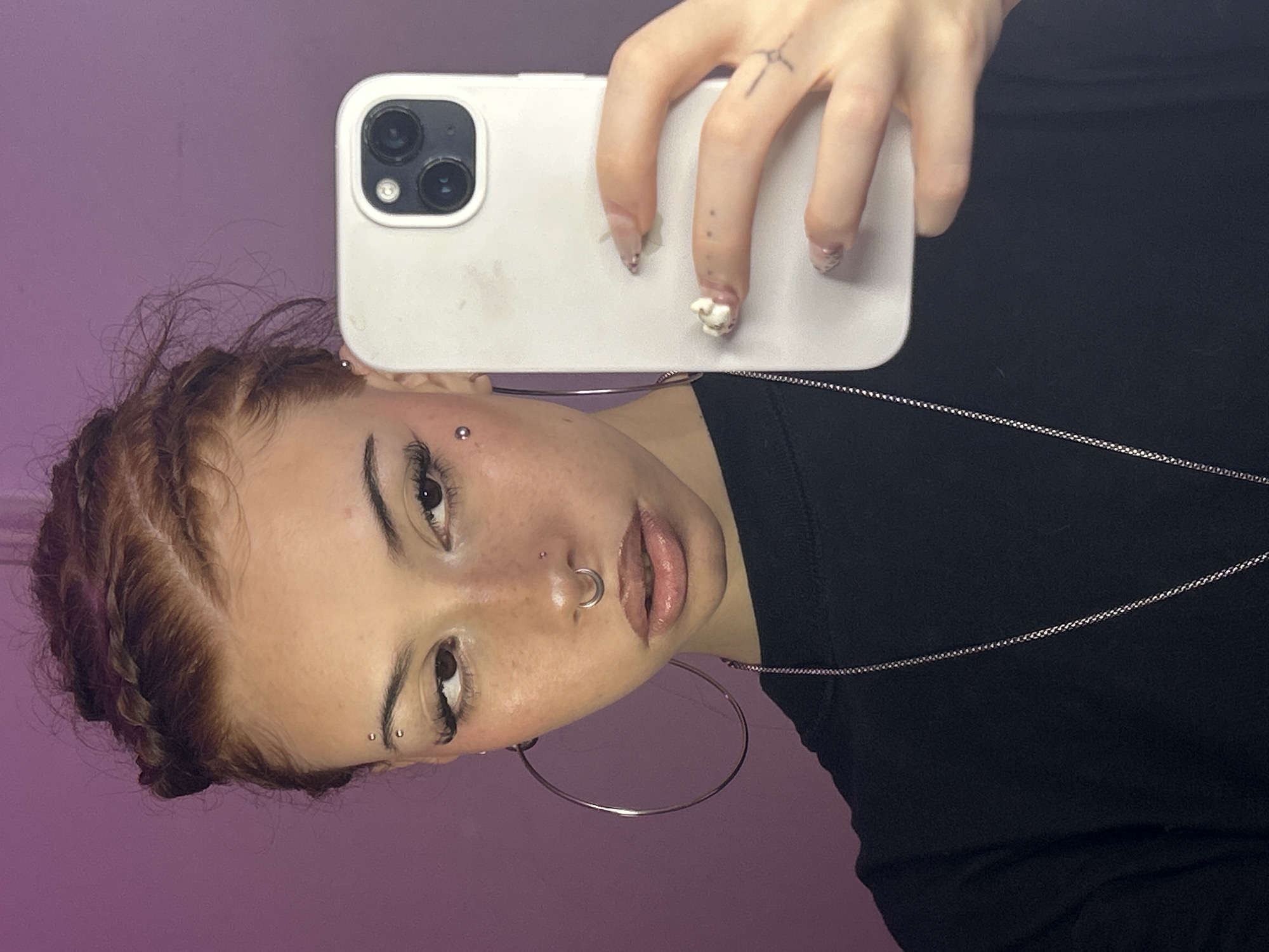 A person with braided red-brown hair, dark eyes, and fair skin is holding a white smartphone in their right hand, taking a selfie against a purple background. The person has multiple facial piercings, including eyebrow piercings, a nose ring, and a piercing near the eye. They are wearing a black top and a silver necklace, and large hoop earrings. The person has distinct nail art on their right hand, with a small embellishment on one nail. The location is not evident from the provided image.