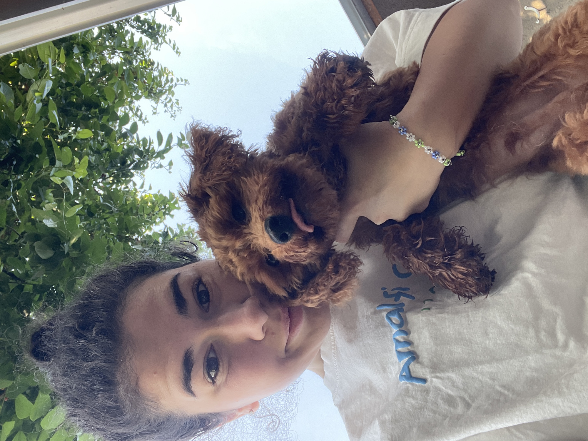 A woman with dark hair is holding a brown dog. The dog is sticking its tongue out. The woman is wearing a white t-shirt and a beaded bracelet. Green trees are visible in the background. The location cannot be determined to be Sofia, Bulgaria from the image alone.