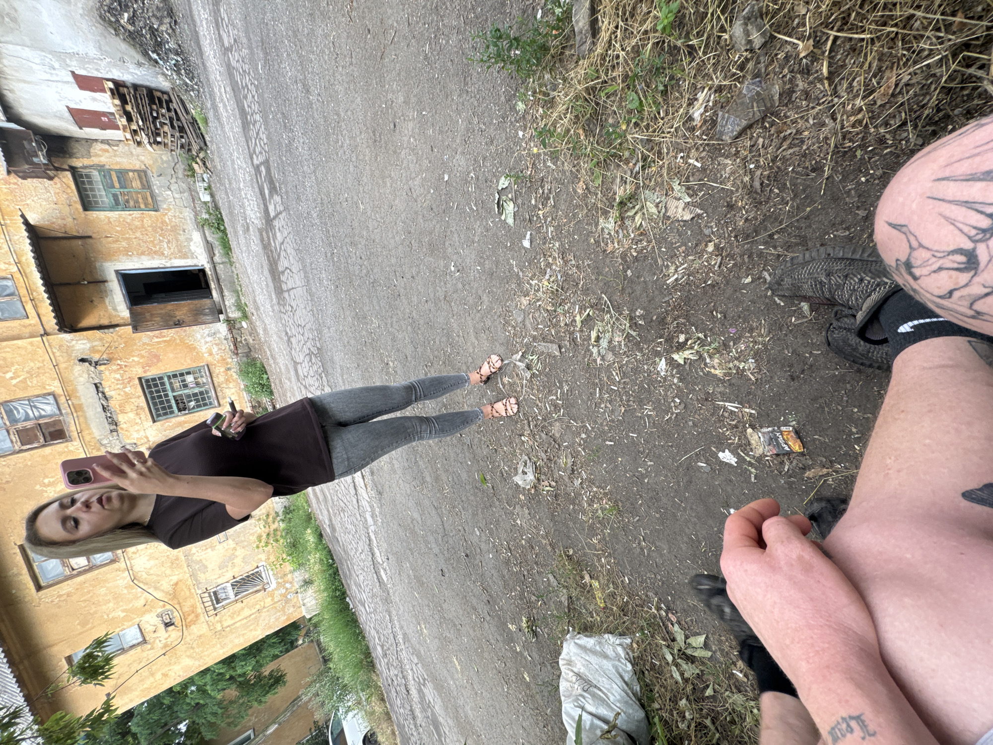 A woman with blonde hair stands in front of a weathered, yellow building, holding a pink smartphone and what appears to be a vape. She is wearing a dark t-shirt and grey jeans, with patterned sandals. Her stance and the phone's position suggest she is taking a selfie or recording. The building behind her has a rustic appearance with visible decay, including peeling paint and a slightly tilted perspective of the structure. The ground is a rough, cracked asphalt surface, littered with debris such as dried leaves, plastic wrappers, and a discarded cigarette pack. To the left of the woman, a white car is partially visible. In the foreground, the legs of a person are visible, one with a tattoo on the thigh, suggesting they are seated or lying down. This person is wearing dark shoes and socks. The overall environment appears to be an urban or semi-urban setting, possibly an alleyway or a neglected courtyard, characterized by its dilapidated structures and unkempt surroundings.