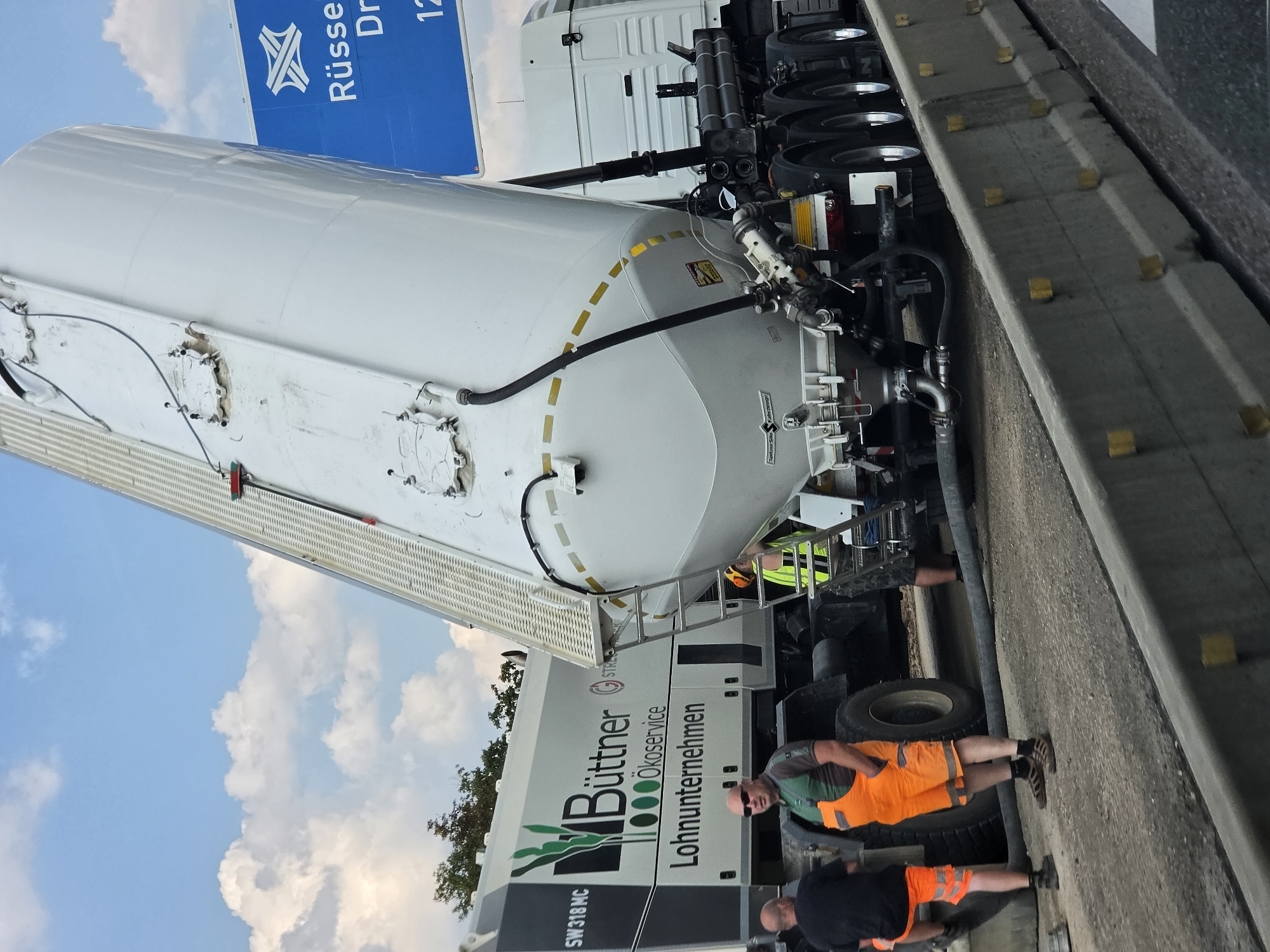 The image captures a scene on the B42 highway near Rüsselsheim am Main, Germany, where a large white cement mixer truck with a tilted silo is parked on the roadway. The truck bears the branding "Büttner Ökoservice Lohnunternehmen" and "SW 318 MC". Two men in work attire are present. One man, wearing orange overalls and a green shirt, stands facing the camera. Another man, in a black t-shirt and orange shorts, is partially visible to the left, leaning against the truck. A third person in a yellow vest is also visible near the rear of the cement mixer. The truck's silo is elevated, suggesting it is in the process of unloading or loading. The surrounding environment includes a concrete barrier separating the roadway and a blue highway sign indicating directions to Rüsselsheim and Darmstadt. The sky is partly cloudy.