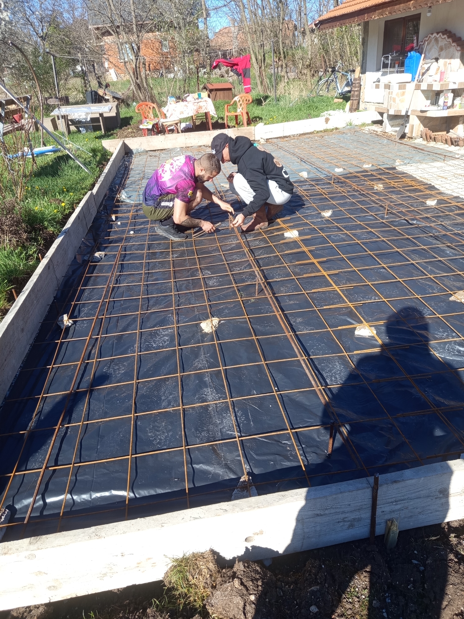 Two men are tying rebar together on a construction site, in the city of Stefanovo, Bulgaria. The site is a rectangular wooden frame with a black plastic liner. On top of the liner, a grid of rebar is being tied together by the men. One man is wearing a purple shirt, and the other is wearing a black hoodie. The men are squatting and using pliers to tie the rebar. The background shows a yard with tables, chairs, a house, and trees. The shadow of a person is visible in the foreground.