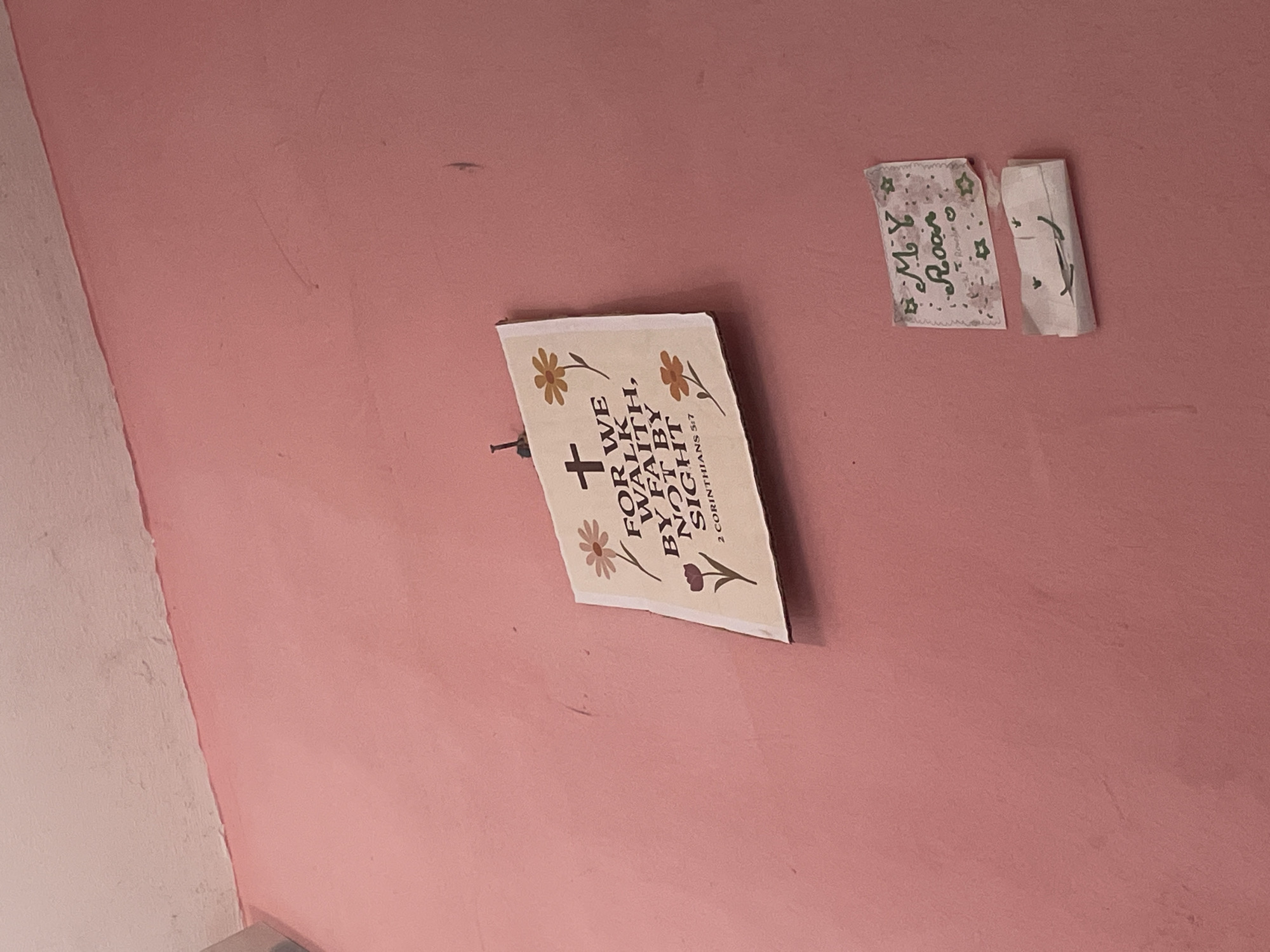 The image shows a pink wall in a room. Two pieces of paper are affixed to the wall. The upper paper features a Bible verse: "FOR WE WALK BY FAITH, NOT BY SIGHT, 2 CORINTHIANS 5:7." It is decorated with drawings of flowers. Below this, a smaller piece of paper displays a drawing of a smiley face. The room appears to be located in Iyafin, Nigeria. The wall is painted a light pink color, and the ceiling above is a lighter shade of pink and white. The items are mounted with what appear to be small nails.