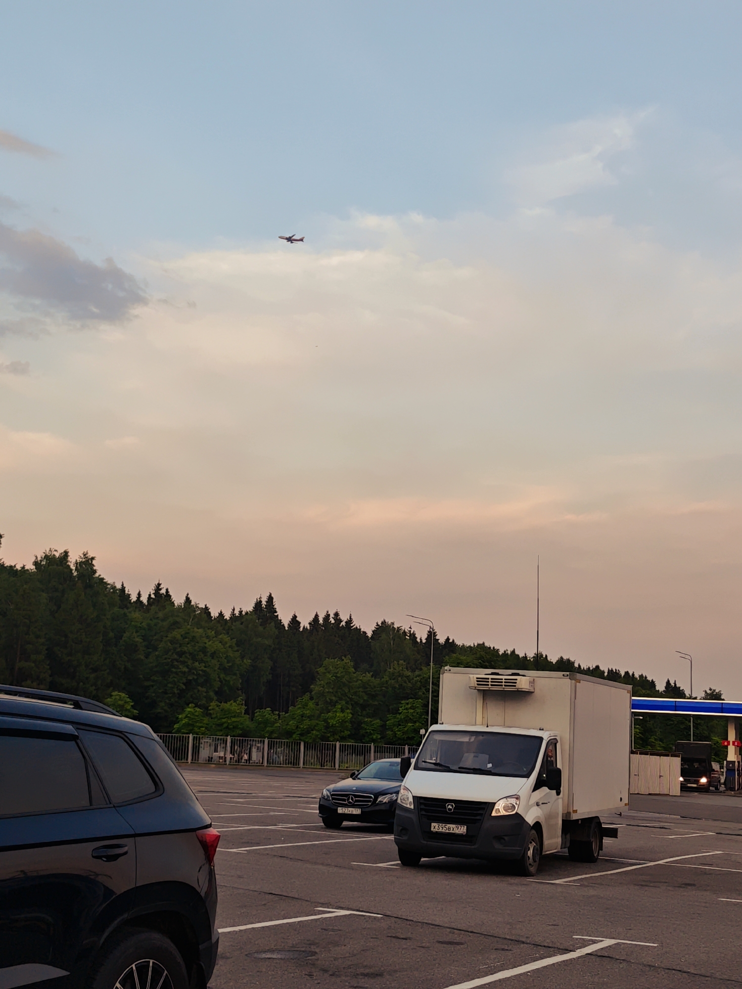 The image captures a scene on a late afternoon or early evening, with a pale pink and blue sky filled with scattered clouds. An airplane is visible in the upper left portion of the sky. In the foreground, a dark-colored SUV is parked on the left side of a parking lot, its rear visible. Further into the parking lot, a white refrigerated truck is driving forward, its headlights on. To the left of the truck, a black Mercedes sedan is parked. In the background, a dense line of dark green trees forms a forest canopy. Beyond the trees, a gas station with a blue canopy and visible pumps is situated on the right. Streetlights are present in the middle ground. The scene is set in the city of Sholokhovo, Russia, likely at a gas station or rest stop adjacent to a wooded area.
