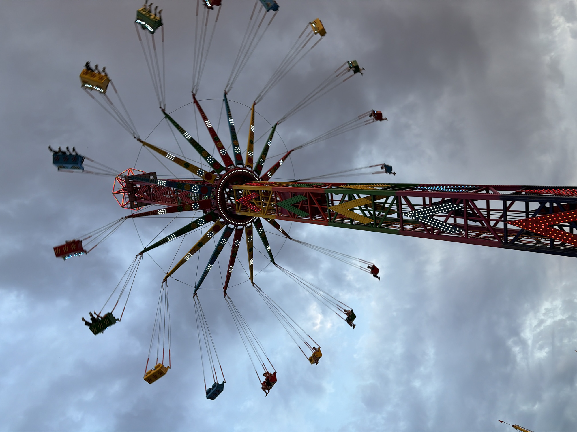 A close-up, low-angle shot captures a colorful swing ride at a fair or amusement park against a cloudy, dusky sky. The central tower of the ride is tall and segmented, with sections of red, yellow, green, and blue supports. Strings of lights adorn the structure, some blinking white, others red. Extending outwards from the tower are numerous arms, each supporting a swing. These arms are also decorated with colorful lights, appearing as red, yellow, blue, and green segments. Several swings are visible, each occupied by one or two individuals. The swings are in motion, giving a sense of activity and the ride's rotation. The sky above is a mix of dark gray and lighter blue clouds, with a hint of fading daylight. The overall scene conveys a sense of evening amusement and excitement.