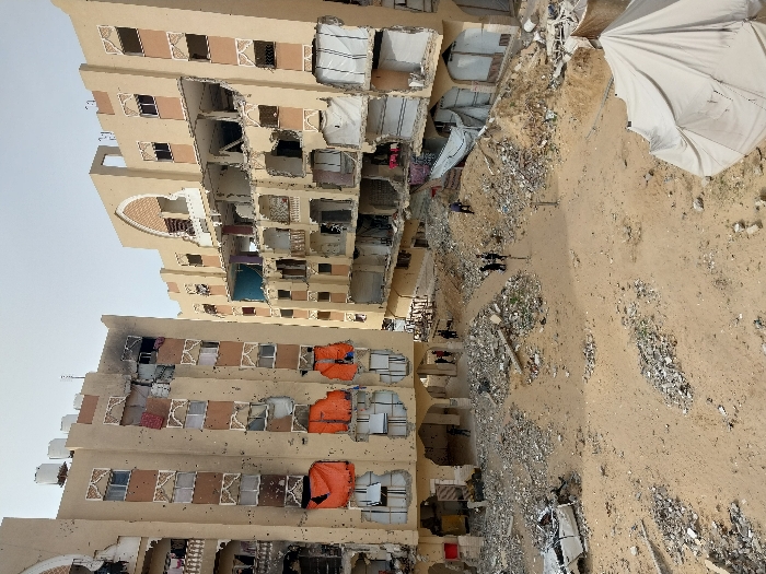 The image depicts a devastated urban scene in Khan Yunis, Palestinian Territory, characterized by extensive structural damage to residential buildings and debris-strewn ground. Two multi-story, light-colored apartment buildings dominate the background, both showing severe damage. Their facades are riddled with numerous circular impact marks, consistent with bullet holes or shrapnel. Many windows are shattered, missing, or covered with makeshift materials such as white sheets and orange tarpaulins. The left building exhibits burn damage near its upper-middle section, and several white water tanks are visible on its roof. The right building has significant structural damage, with entire sections of walls and balconies missing, exposing internal floor plans and twisted rebar.
The ground in the foreground and midground is composed of disturbed sand and earth, heavily scattered with large piles of concrete rubble, twisted metal, and various debris. A partially destroyed white vehicle, likely a car, lies on its side in the mid-left foreground. In the lower right corner, a light-colored canvas tent is partially visible. Several individuals are present within the scene: a group of approximately 3-4 people stands in the center-midground amidst the rubble, and other individuals are seen near the ground floors and under the archways of the damaged buildings. The sky is overcast or hazy, providing diffused natural light.