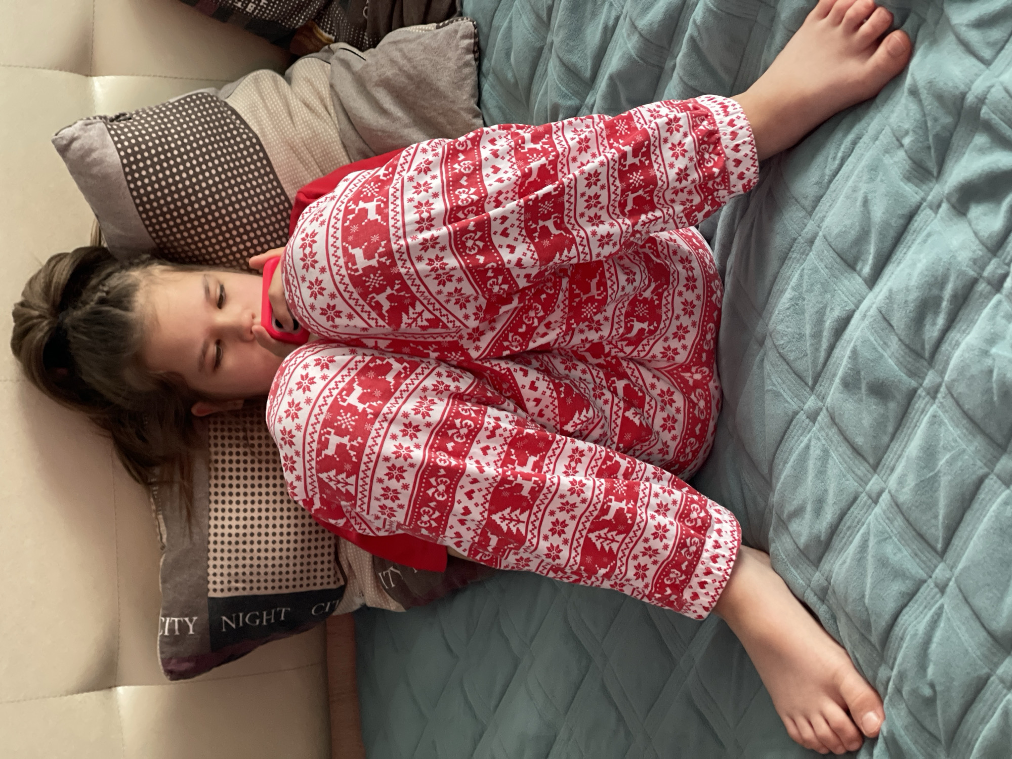 A young girl is depicted sleeping or resting indoors, curled up on what appears to be a light-colored couch or bed. She is lying on her left side, with her head resting on a grey pillow that has a pattern of white dots and the text "NIGHT CITY" visible on its edge. Her brown hair is tied up in a high ponytail or bun. She is wearing red and white pajamas adorned with a festive pattern featuring reindeer and snowflakes, suggesting a holiday or winter theme. A small pink object, possibly a pacifier or phone, is held near her chin. Her bare feet are visible, resting on a textured, light blue or teal quilted blanket that covers the surface beside her. Another plain grey pillow is partially visible behind her head. The overall scene conveys a sense of peaceful slumber or relaxation.