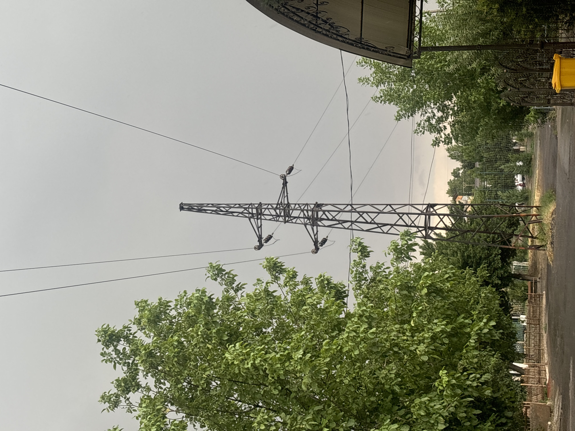 The image depicts an outdoor scene with an overcast sky. A tall, lattice-style power pylon stands prominently in the center, supporting thick electrical cables that stretch across the frame. To the left, a large, leafy green tree dominates the foreground, its branches reaching towards the sky. In the background, a dense line of trees and greenery forms a natural barrier. A portion of an arched structure with decorative metalwork is visible on the right side of the image, casting a shadow. The ground appears to be a dark, paved surface, possibly a road or pathway, with some gravel and grass visible at the edges. A yellow trash bin is situated near the right edge of the frame, beside a black metal fence. The overall setting suggests a semi-urban or suburban area.