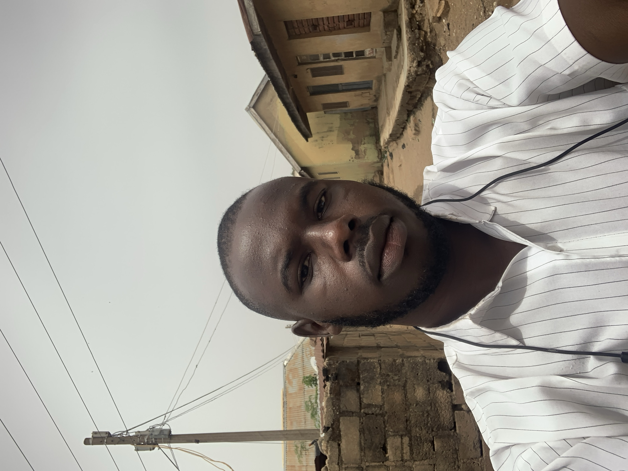A medium, outdoor, daytime shot depicts a man in a white, pinstriped shirt. A black wire, likely from headphones, is visible around his neck. He has a dark complexion, a short beard, and closely cropped hair. Behind him are structures indicative of a developing area. There's a building with a dilapidated facade and exposed brickwork. In the background are utility poles with wires, suggesting an urban or semi-urban location. Given the reference to Jos, Nigeria, the surroundings appear to be consistent with the infrastructure and architecture of a residential or commercial neighborhood in that region. The sky is overcast.