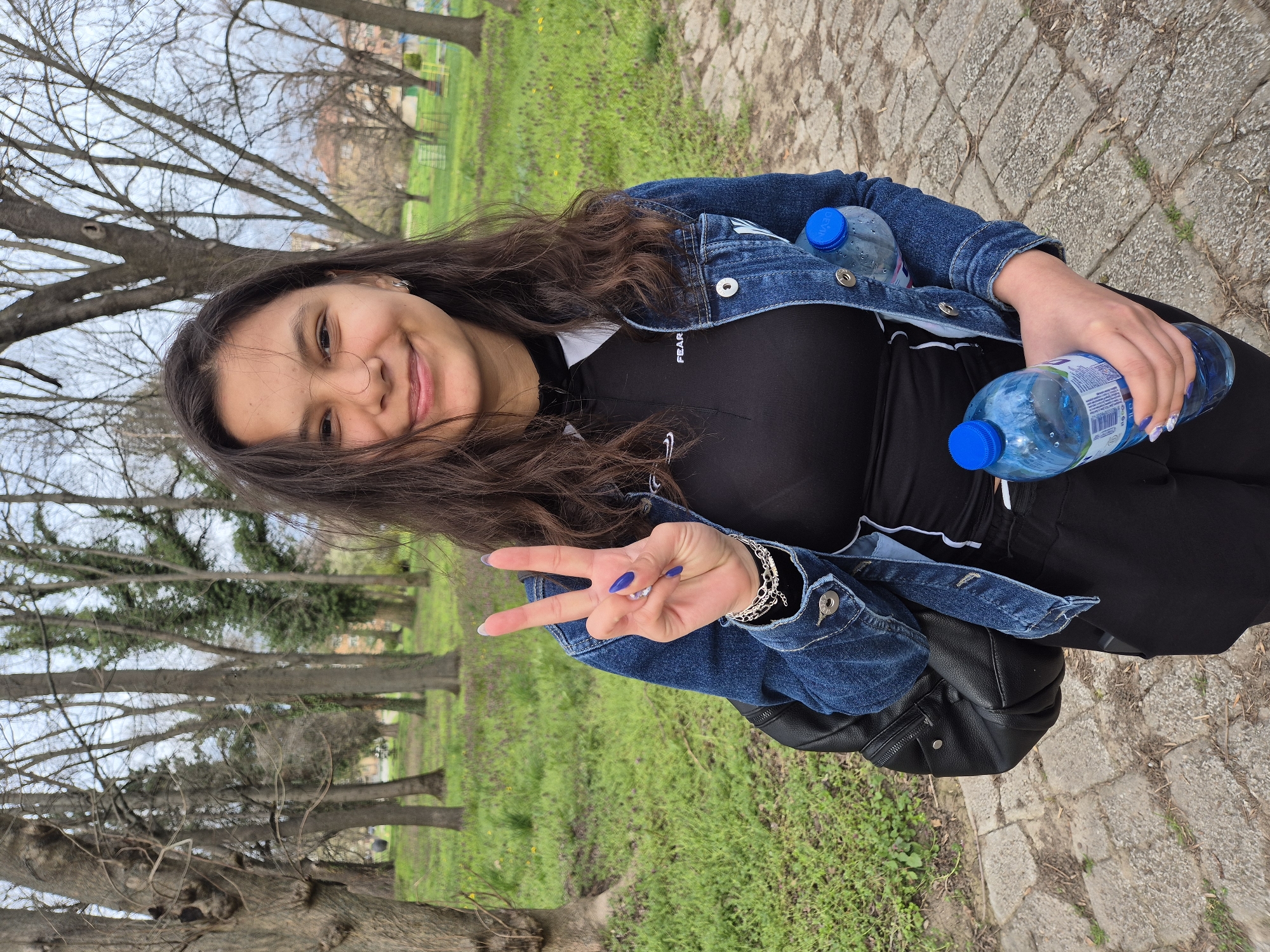 A young woman stands outside in what appears to be a park. She is holding two bottles of water, and making a peace sign with her hand. She is wearing a denim jacket, black shirt, and black pants. In the background, there are trees and green grass. The surface beneath her feet is a paved walkway. The location is Pavlikeni, Bulgaria.