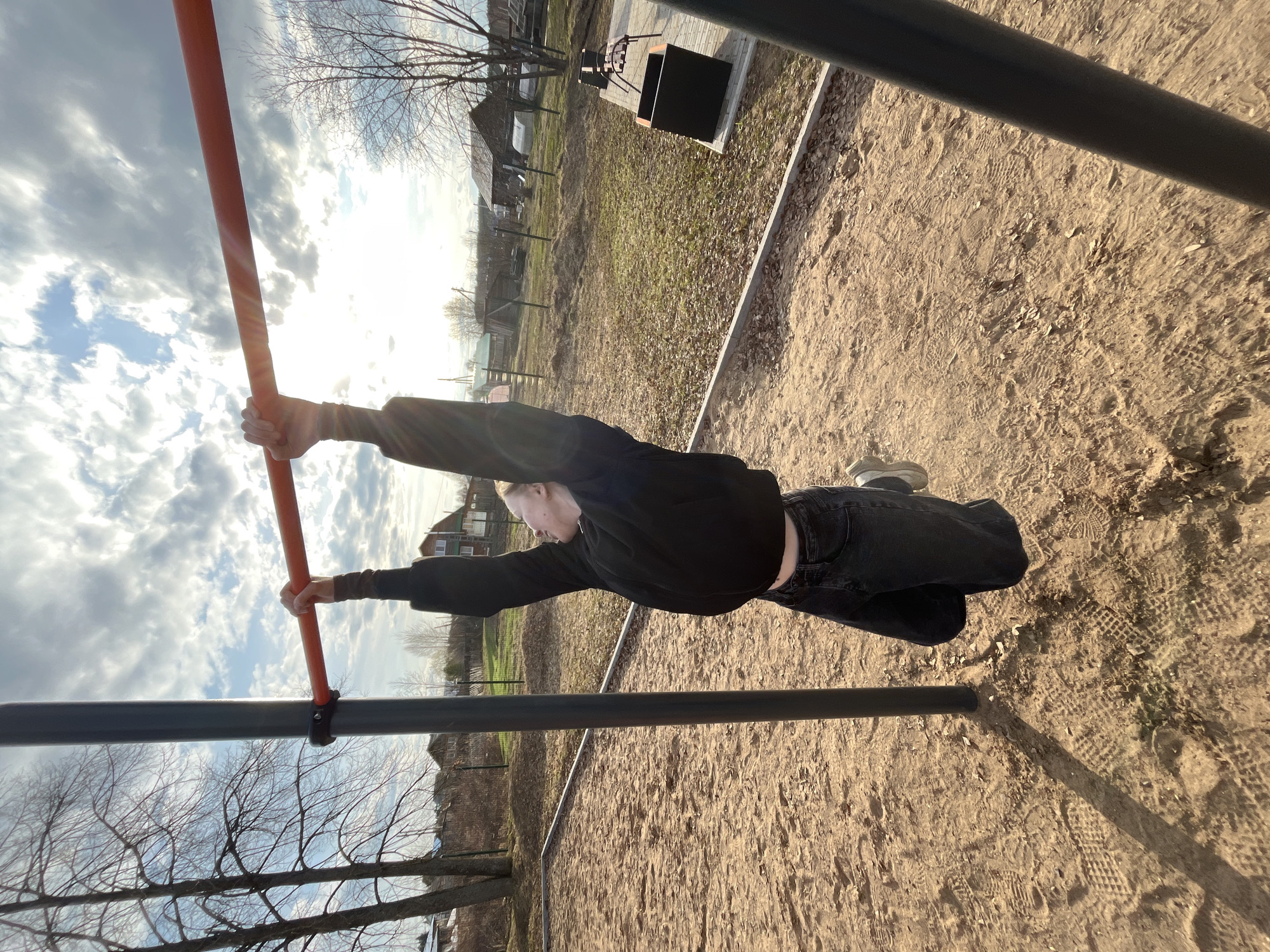 The image captures a young individual performing an exercise on a horizontal bar in an outdoor setting in Сосново, Russia. The subject, dressed in a black long-sleeved top and dark jeans, is seen hanging from a bright orange/red horizontal bar. Their body is stretched out almost parallel to the ground, arms fully extended overhead gripping the bar, indicating a calisthenics move or an advanced stretch. Another darker, possibly black or grey, horizontal bar is positioned lower down.
The scene takes place on what appears to be a sandy or dirt ground, marked with numerous footprints and scattered dry leaves. In the background, there are bare trees, suggesting the season is late autumn, winter, or early spring. Several distant houses or buildings are visible through the sparse tree line, hinting at a semi-rural or suburban environment. A small paved area in the background features a black metal structure, possibly a grill or a waste container.
The time of day is clearly sunny, with bright sunlight streaming from the upper right, illuminating the scene and creating a warm ambiance. The sky is partly cloudy, with patches of blue visible amidst white and grey clouds. No text is visible in the image. The overall impression is one of focused physical activity in a simple, outdoor fitness area.