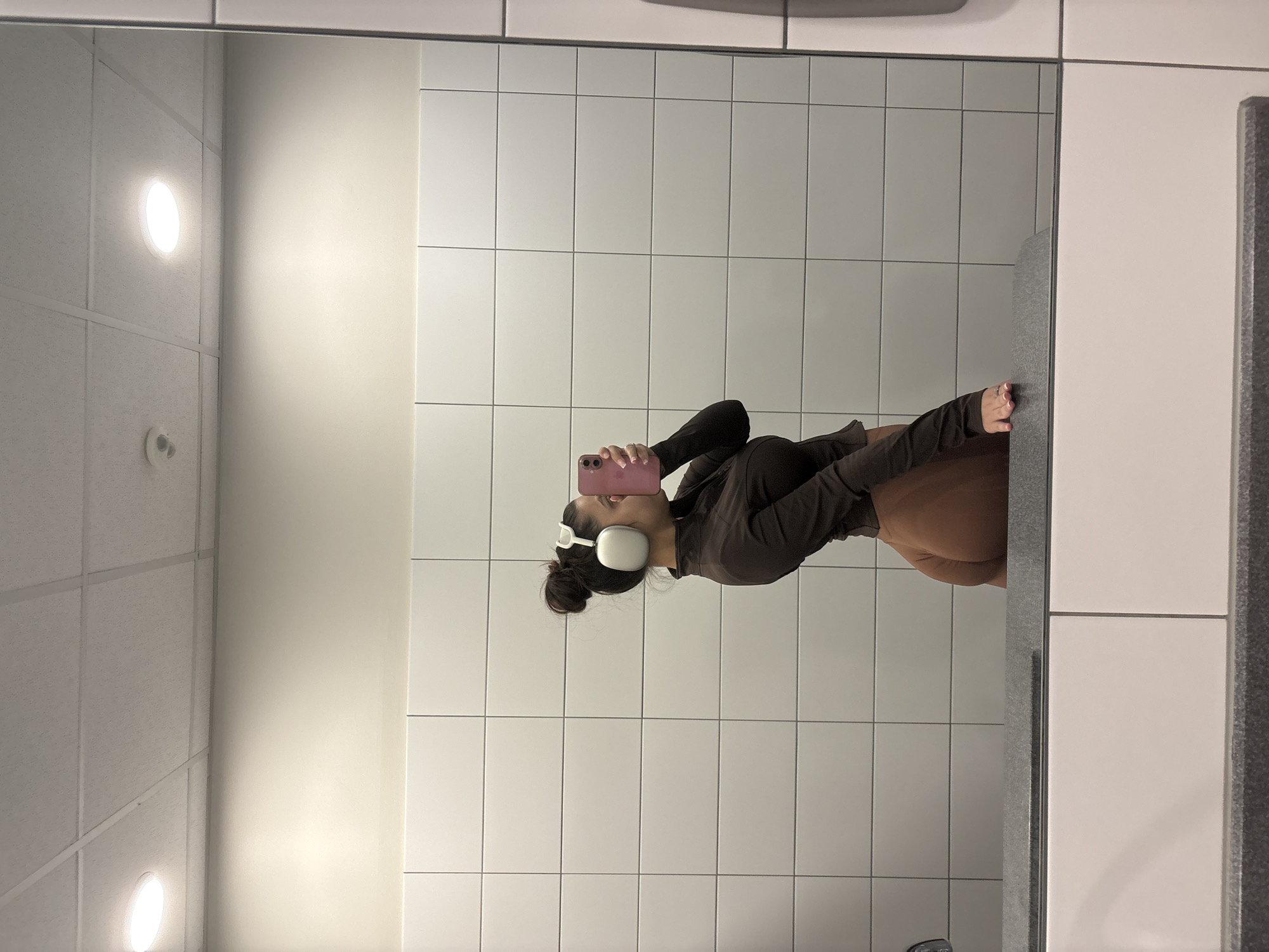 A woman is reflected in a mirror, holding a pink phone up to her face, wearing white headphones, and a brown long-sleeved shirt. She is in a bathroom with white tiles and a white ceiling with recessed lighting. The location is unknown and cannot be determined from the given image.