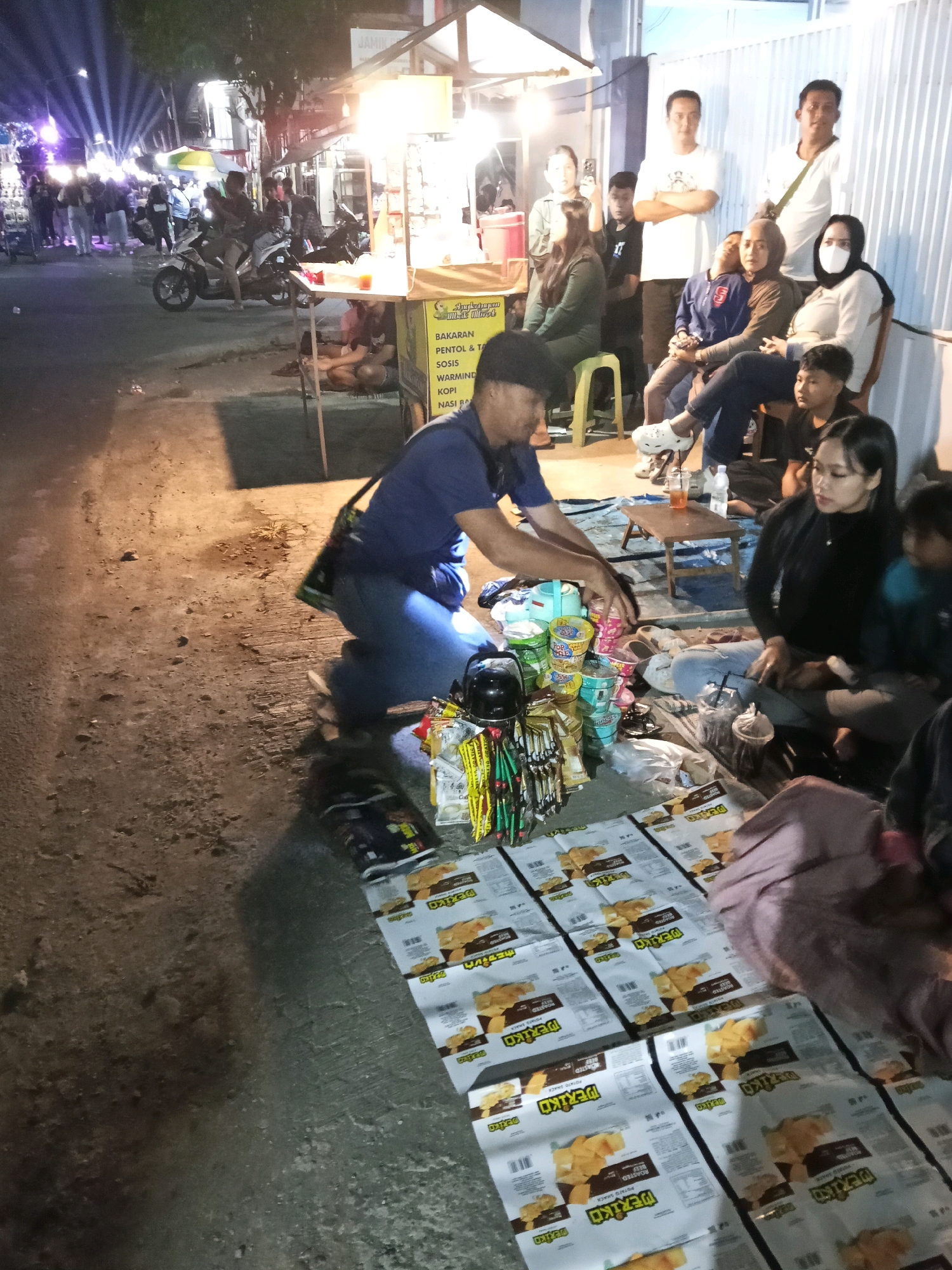 The image depicts an outdoor scene at night, likely a street market or event, in Bendosewu, Indonesia. In the foreground, several people are gathered on the ground, seated on a tarp. A man in a blue shirt and blue jeans is kneeling, arranging what appear to be snacks and beverages. In front of him are various food items, including cup noodles, plastic bags of chips, and what seem to be packaged snacks. To his right, a woman with long dark hair is seated and looking towards the man. Other individuals are present, some standing and some seated in the background.
The environment suggests a temporary setup. There are food stalls visible in the mid-ground, illuminated by bright lights, with a motorcycle parked nearby. In the background, more people are visible, along with lights that suggest a gathering or performance. The ground is paved with asphalt, and there are shadows cast by the artificial lighting. The overall atmosphere appears to be that of a bustling night market.