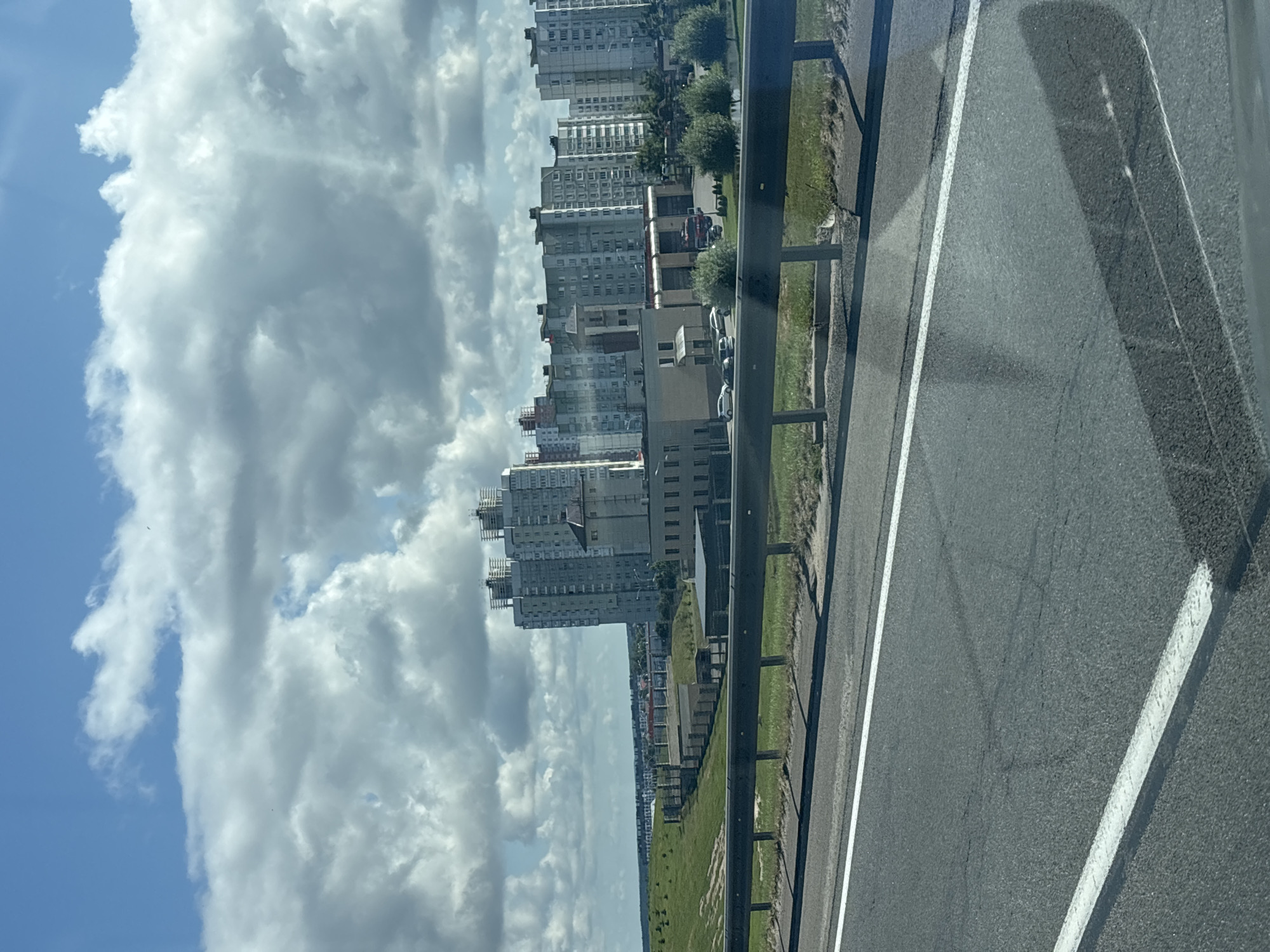 A photograph captures a view of apartment buildings and a fire station from a highway in Minsk, Belarus. The sky is partly cloudy with large white cumulus clouds against a blue sky. In the foreground, a section of the highway is visible with a black metal guardrail running along its edge. Beyond the guardrail, a green grassy hill slopes upwards towards a line of buildings. These buildings include a cluster of modern, tall apartment buildings, some appearing under construction. In front of the apartment buildings, a lower-rise building that resembles a fire station with red doors is visible. Several cars are parked in front of the fire station. The overall impression is an urban fringe landscape.
