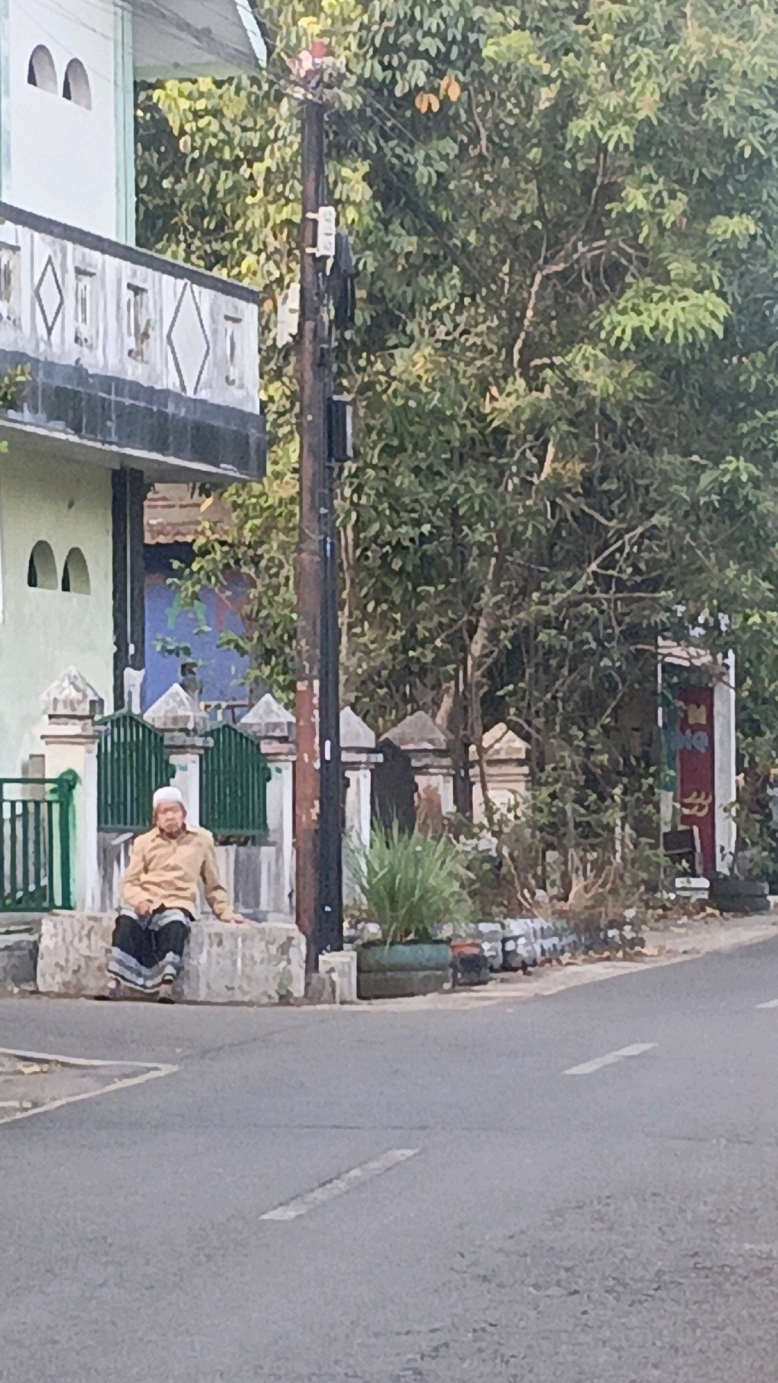 In Salatiga, Indonesia, a man wearing a white cap and a light brown long-sleeved shirt is seated on a low concrete wall outside a building. He is also wearing a dark sarong. The man is facing left, with his hands resting on his lap. The building behind him has a light green facade and decorative white elements. A dark green metal fence is positioned in front of the building. A wooden utility pole with electrical wires and various attachments stands to the right of the man and the building. Lush green foliage from trees and shrubs surrounds the area, creating a natural backdrop. The scene is set on a paved road with a faint white line marking the lane. The lighting suggests it is daytime, possibly late afternoon given the warm tones of the man's shirt.