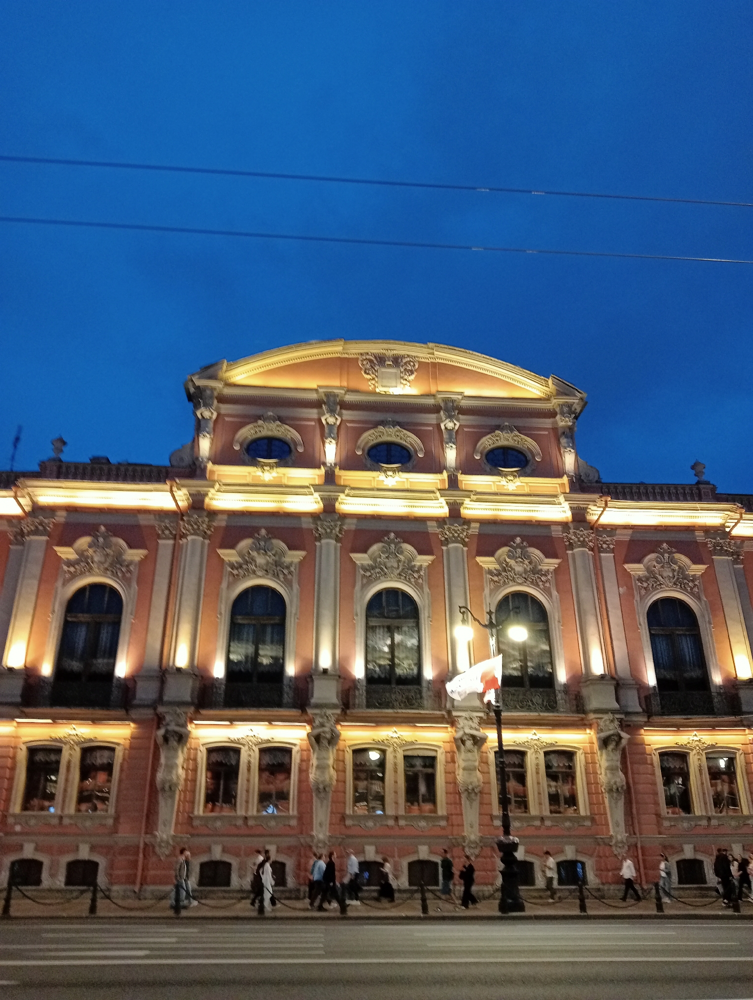 The image depicts the exterior of the Stroganov Palace in Saint Petersburg, Russia, during the evening twilight. The palace is illuminated by warm artificial lights, highlighting its ornate Baroque architecture, characterized by elaborate carvings, columns, and arched windows. Two rows of windows are visible on the facade, with the upper row featuring oval windows. A flag, white with a red stripe, flutters from a lamppost in front of the building. Several people are walking on the sidewalk in front of the palace, with some crossing a crosswalk on the street. The sky above is a deep blue, with two distinct horizontal power lines stretching across it. The overall scene captures a moment of civic life in the city at dusk.