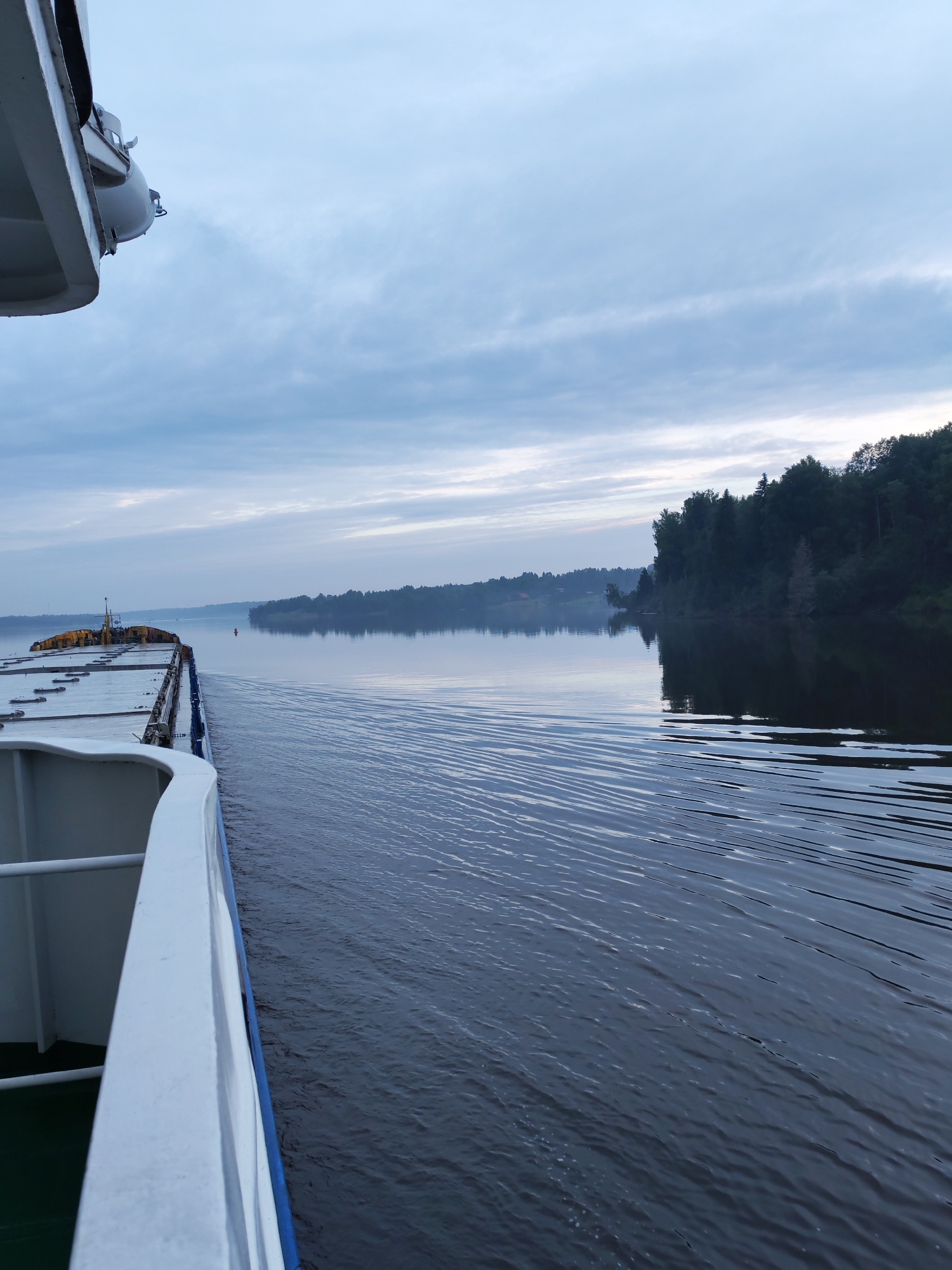 This image depicts a side view from a moving boat on a body of water, likely a river, on an overcast day. In the foreground, the white railing and deck of the boat are visible. The water is dark and shows ripples caused by the boat's movement, with a wake extending behind it. In the distance, a densely wooded shoreline lines the opposite side of the water. The sky is filled with clouds, with a lighter area near the horizon suggesting either dawn or dusk. There are no people visible in the image. The scene is likely located on a river near the village of Kondratovo, Russia, given the provided location context.