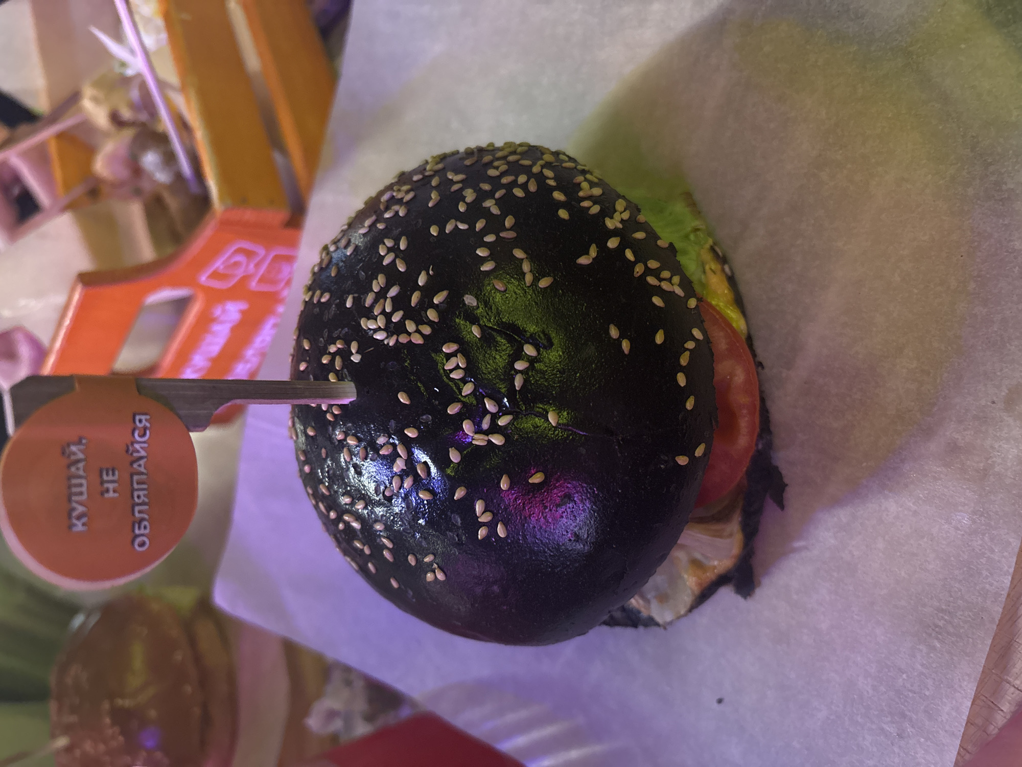 A top-down shot shows a black burger topped with sesame seeds. The burger is pinned with a small orange round sign bearing a Russian inscription. The burger is sitting on white parchment paper. The setting is likely in a restaurant. Based on the text on the sign, the location is likely in the city of Saint Petersburg, Russia.