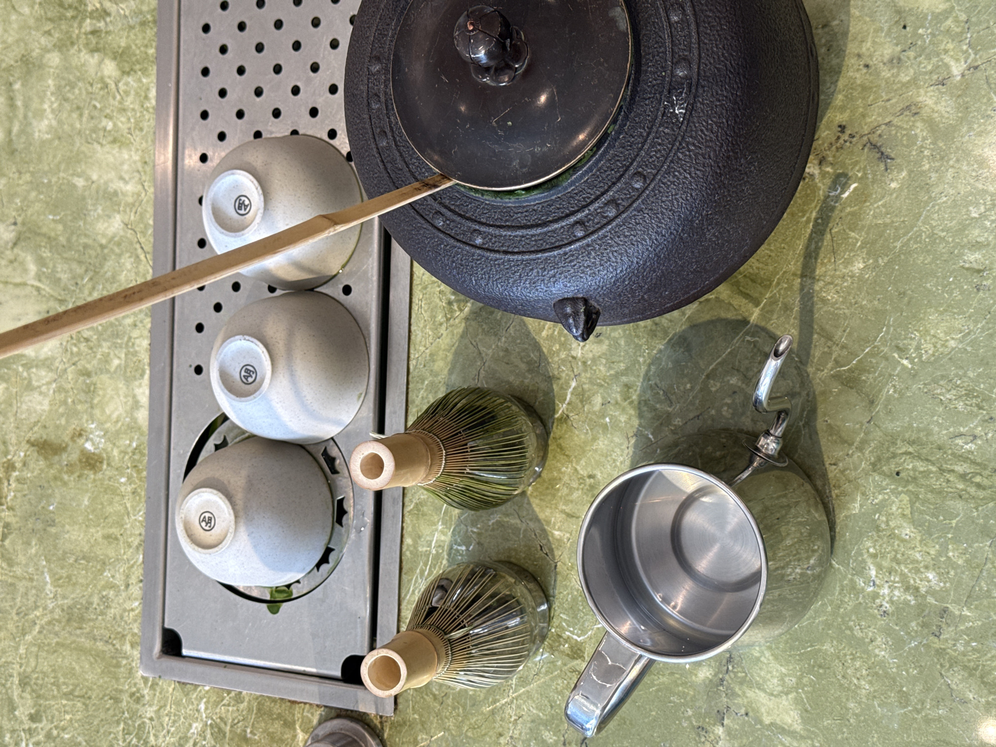 The image shows a tea preparation setup on a green, possibly marble, surface. A dark, textured teapot sits to the right, with a bamboo utensil extending from it towards a metal tray containing three small, light-colored bowls. Two green, whisk-like objects are positioned below the teapot and adjacent to a stainless steel pitcher. The scene is located in Beverly Hills, United States of America.