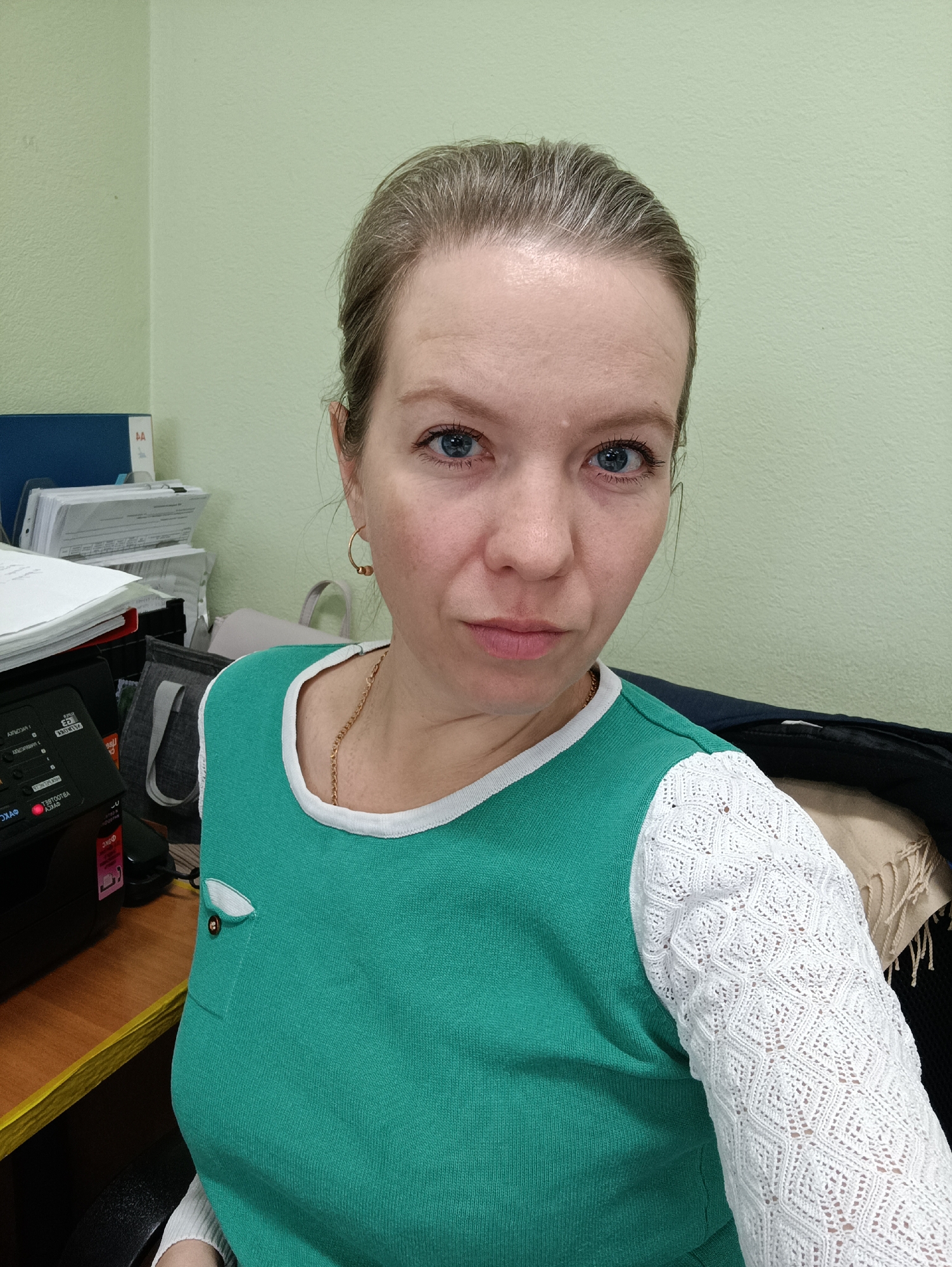 A medium close-up selfie captures a woman looking directly at the camera, seated indoors at what appears to be a desk in an office environment in Chaikovsky, Russia. The woman has light skin, blue eyes, and light brown hair pulled back, with some grey strands visible near her forehead. She is wearing a teal green top with a white trim around the neck and distinctive white lace sleeves with a geometric pattern. A thin gold chain is visible around her neck, and she wears small gold hoop earrings. Her expression is neutral to slightly serious.
The background shows a light green wall. To her left, an office desk is cluttered with stacks of papers, a blue folder marked "4A," and a black piece of office equipment (possibly a printer or scanner) with various Cyrillic labels, some appearing mirrored, such as "АВДО...Т", "ФКС", and "ТВТ...К". A grey bag is partially visible behind the papers. To her right, part of a darker piece of clothing or fabric, possibly a jacket or blanket, is visible. The lighting suggests it is daytime.