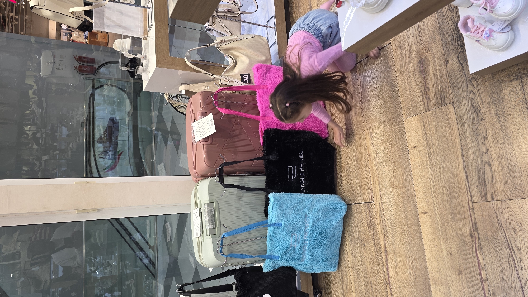 The image shows an indoor scene, likely a retail store, as suggested by the display of merchandise. A young girl with long dark hair, dressed in a pink shirt and a blue denim skirt, is reaching toward something. Next to her is a display of luggage and handbags. The stack of luggage includes a rose-colored suitcase, a light-green suitcase, a fuzzy black bag, a fuzzy pink bag, and a fuzzy turquoise bag. The handbags on a shelf above include a tan-colored tote bag. There is also a display of small white shoes with pink accents on a shelf near the girl.
Reflections in the glass window show elements of the exterior environment. Based on visual landmarks, the location is likely within or near Blagoevgrad, Bulgaria.