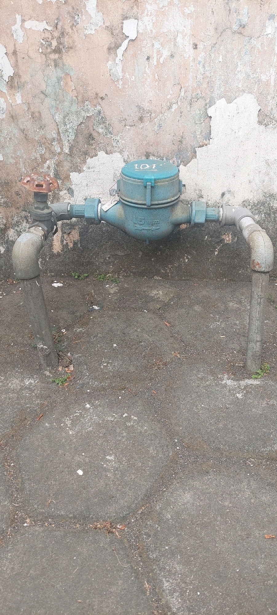 A water meter is installed on a pipe in front of a distressed concrete wall. The water meter is blue and has the number "104" visible on its top. A rusty valve handle is attached to the pipe on the left side of the water meter. The pipe runs horizontally across the frame, supported by two vertical pipe sections. The ground is paved with dark gray concrete slabs and has some scattered debris and small plants. The wall behind the meter is textured and shows signs of peeling paint and discoloration.
