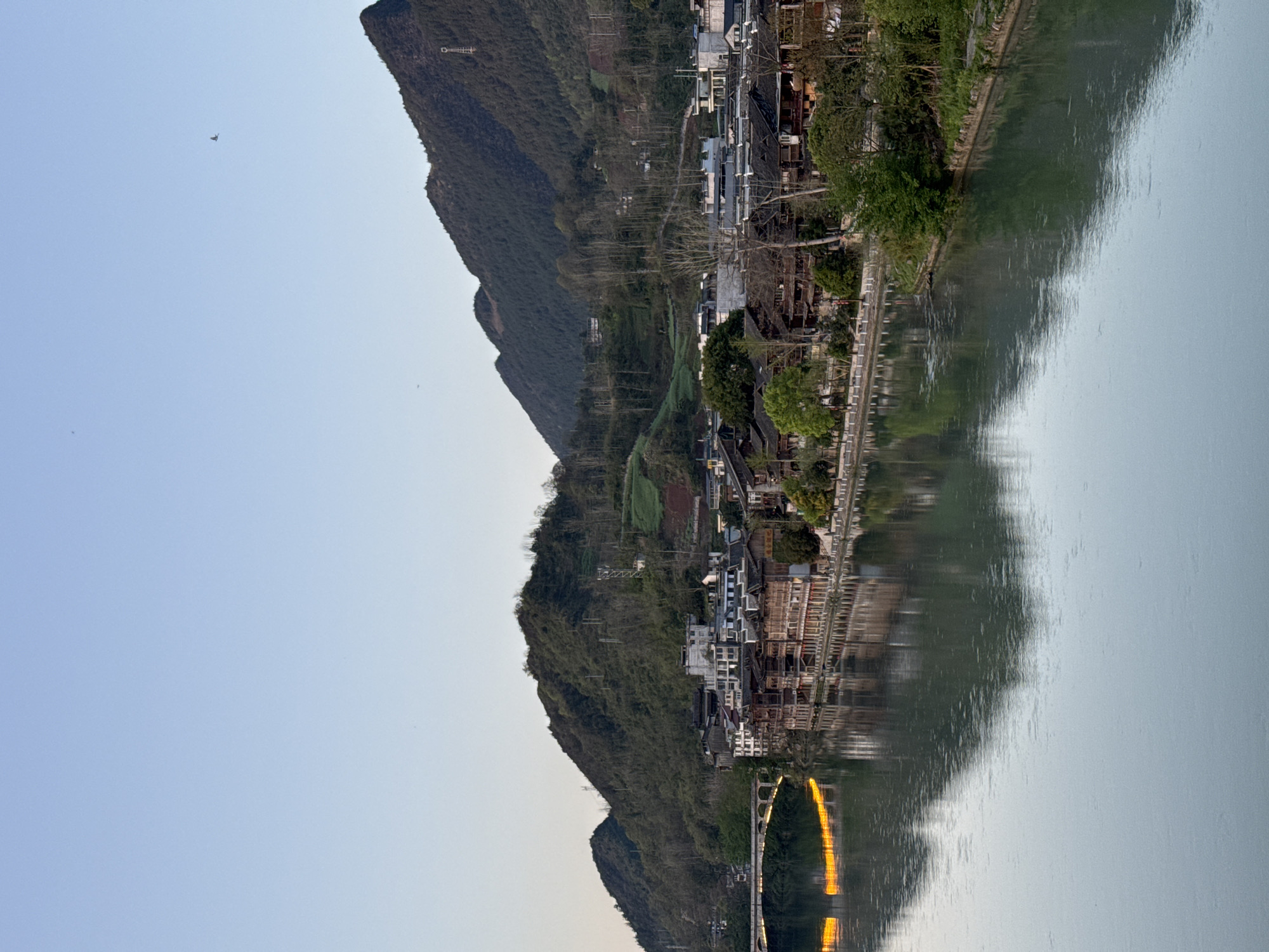 The image depicts a landscape in Zhuoshui, China, featuring a river with reflections of buildings and mountains. A bridge spans the river with warm, yellow light reflecting from its underside. The scene includes mountains covered in vegetation and a settlement with buildings lining the riverbank. The sky is pale blue.