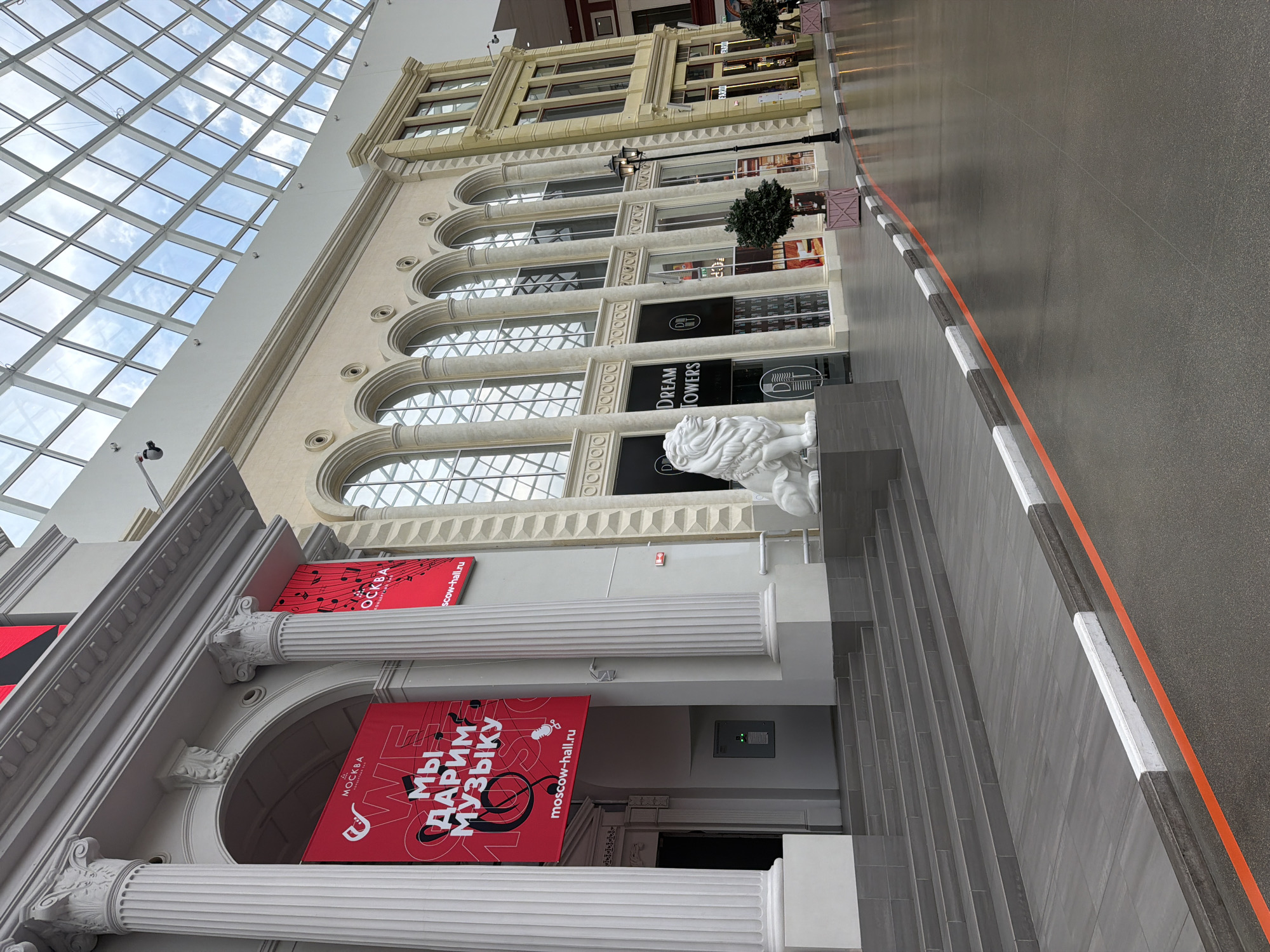 An interior view of a shopping mall with a domed, glass-paneled ceiling. The left side of the image features a classical architectural facade with large white columns and an arched entryway. A red banner with white Cyrillic text and musical notes hangs from the facade. To the right, a cream-colored building with arched windows stretches into the distance, displaying storefronts. In front of the cream building, a white statue of a lion sits on a pedestal. A lamppost stands near the lion. The floor is a polished dark gray material, marked with an orange line and a border of white and gray curbing. Steps lead up to the classical facade on the left. The environment appears to be clean and well-lit, with no people visible.