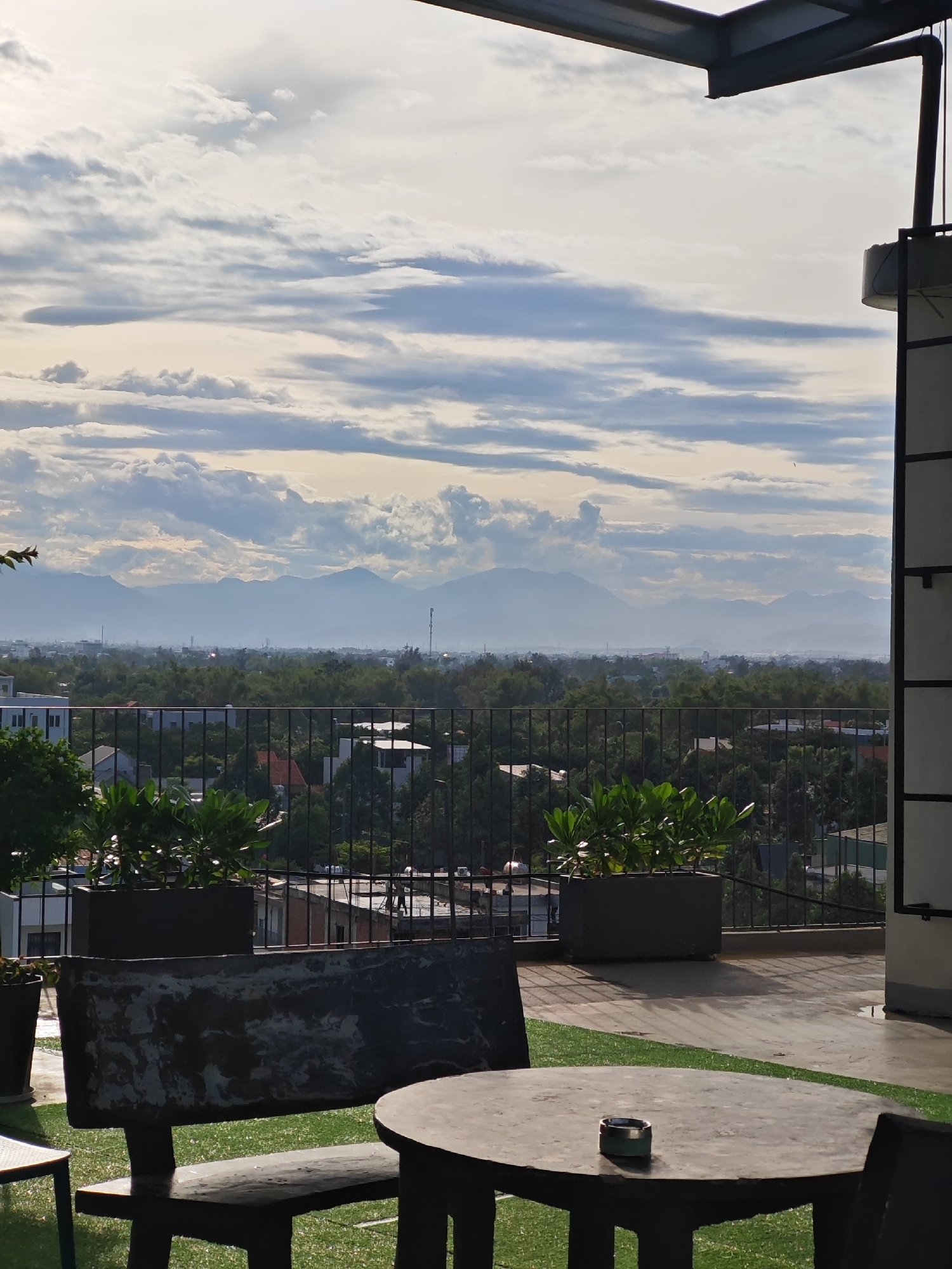 This image depicts a rooftop view in Da Nang, Vietnam, likely from a cafe or restaurant. The foreground features a dark, rustic outdoor table with an ashtray and two chairs, set on a patio with patches of artificial turf. Beyond the patio, a black metal railing encloses the area, with planters containing lush green foliage.
The middle ground offers a panoramic vista of the city, characterized by a dense canopy of green trees interspersed with buildings. The city stretches towards a distant mountain range, which appears hazy due to atmospheric conditions. The sky is filled with dynamic clouds, a mix of white and grey, suggesting either the early morning or late afternoon light. A tall, dark structure, possibly part of the building's architecture or a nearby utility tower, is visible on the right side of the frame. The overall scene evokes a sense of relaxed observation of the urban landscape.