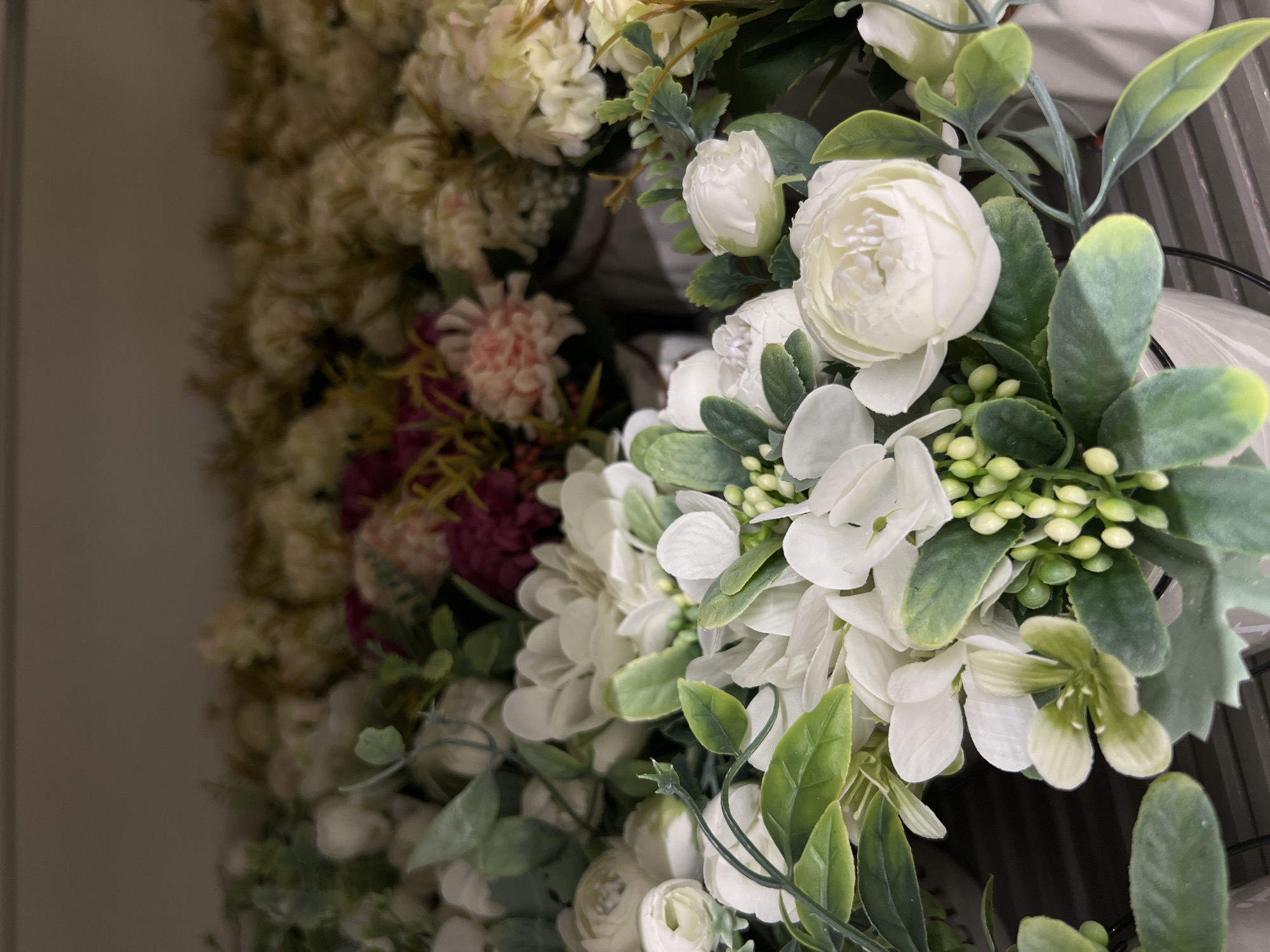 The image displays an arrangement of artificial flowers and greenery. Predominantly, there are white flowers with layered petals, alongside green foliage with rounded leaves. Some of the leaves exhibit a slightly velvety texture. Also present are small, undeveloped buds in a light green color. In the background, a variety of other artificial flowers are visible, including cream-colored and pink blooms. The composition suggests a decorative display, possibly a wreath or floral arrangement. The location in Kazan, Russia is not evident from the visual information provided.