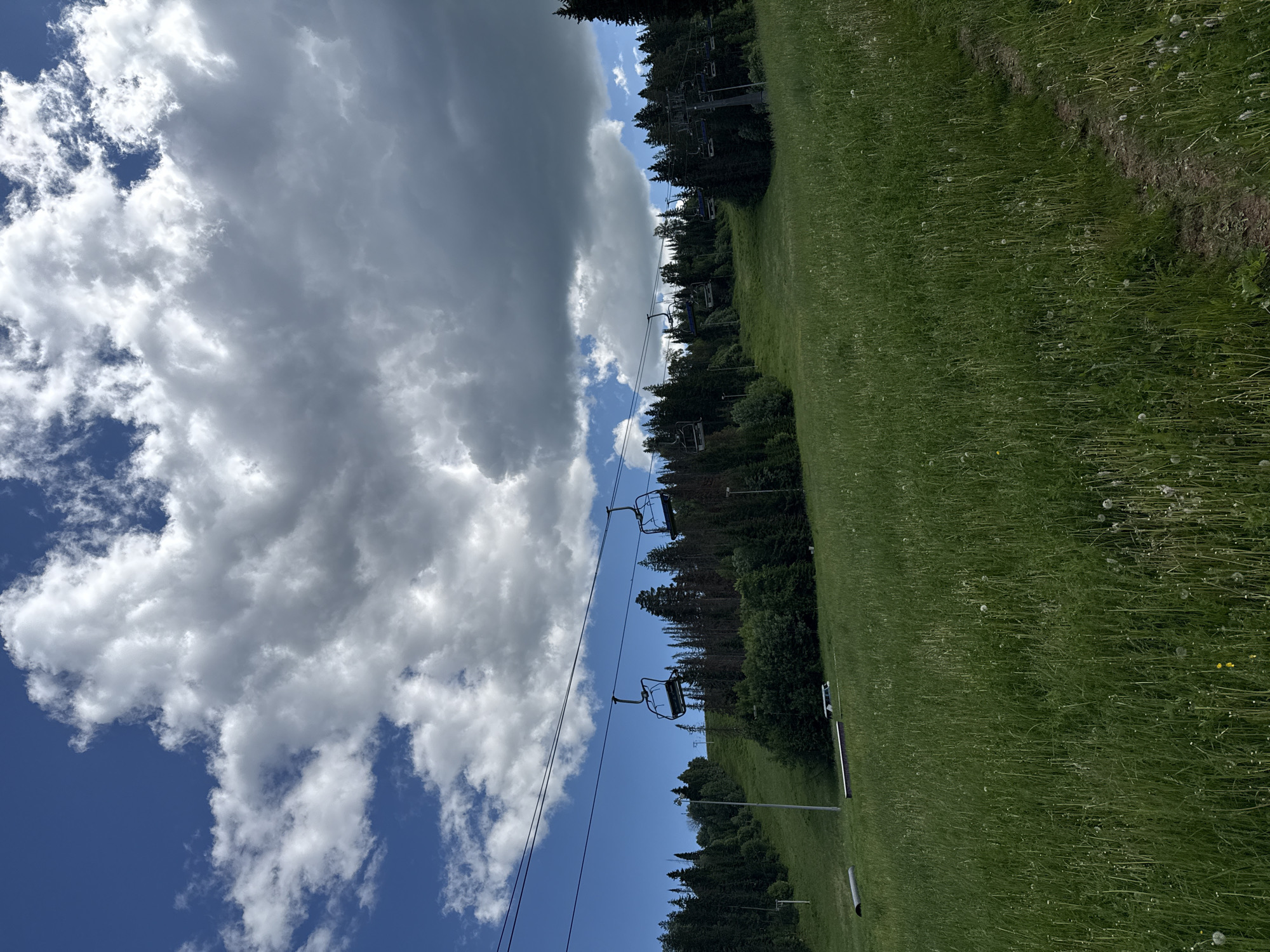 A sunny day with scattered clouds over a green ski slope in Russia. The ski slope is covered in tall grass and wildflowers, with a dirt path winding through it. A ski lift with several chairs is visible in the background, ascending the slope. Trees line the top of the slope, forming a dense forest. The sky is a vibrant blue, with fluffy white clouds adding texture to the scene. A white vehicle is parked on the slope, and a large metal pipe lies on the grass in the foreground.