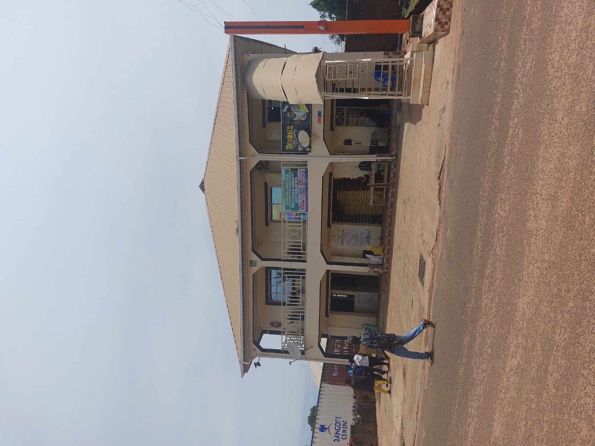 The image shows a two-story building in Barakin, Nigeria, with a light brown, sloped roof. The building is cream-colored with dark trim around windows and doorways. There are balconies on the second floor with white metal railings. Several signs are visible on the building's facade. One sign, partially obscured, appears to be for "SIMON'S" and displays three people's faces. Another sign, on the left, is green and white and reads "PLATEAU STATE FERTILIZER STOCKISTS DISTRIBUTION".
In front of the building, there is a paved area and a road. Several people are present:
- A man in a patterned blue and black shirt and blue jeans is walking across the foreground from left to right.
- A group of three people are standing near a white shipping container on the left side of the image, to the left of the building.
- One person is sitting at a wooden table to the left, in the ground floor of the building.
- Another person is sitting inside the building, near the entrance.
On the left side of the image, a large white shipping container with the text "DANGOTE CEMENT" is visible. There are also stacks of large yellow bags, likely fertilizer, placed outside the building and inside the ground floor openings. The environment appears to be a street or commercial area, with a dirt and paved surface. An orange pole-like structure is visible on the right side of the building. The sky is overcast.