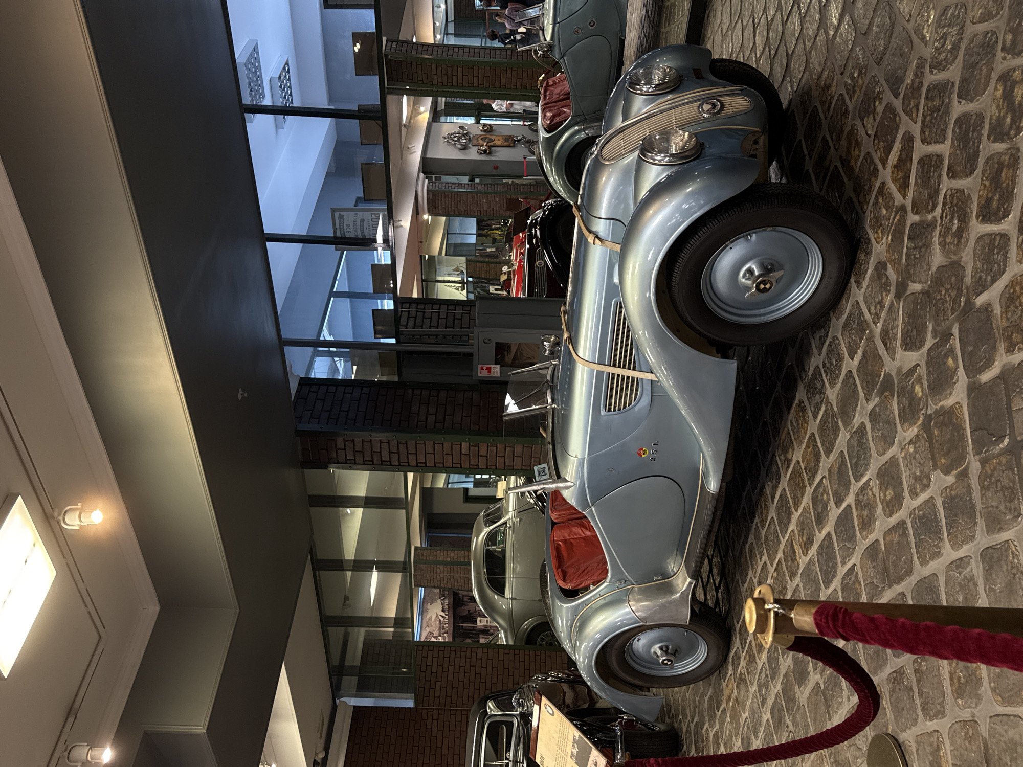 In the museum in Arkhangel'skoe, Russia, an exhibition of vintage cars is on display. The primary focus is on a light blue convertible with red interior seats. The car is positioned on a cobblestone floor, and a red velvet rope on a gold stanchion is visible in the foreground, indicating a display area. Behind the convertible, a silver coupe is visible, and further back, a black car with a display sign. Reflections in the glass walls reveal more exhibits and a well-lit interior space with brick accents on the pillars. The lighting is artificial, coming from ceiling fixtures. No individuals are clearly interacting with the cars; they appear to be static displays.