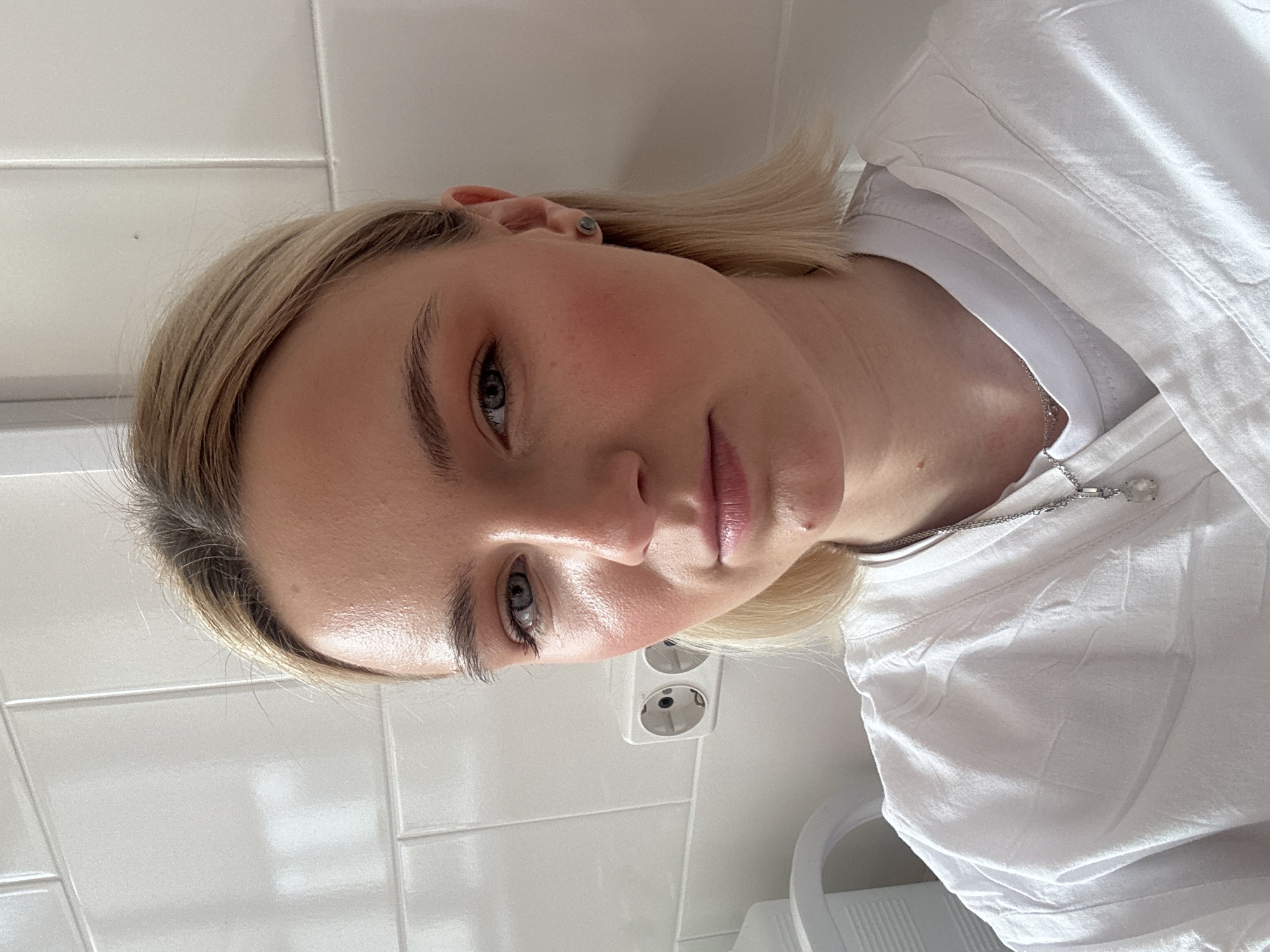 A close-up, slightly rotated image features a fair-skinned individual with blonde hair and light-colored eyes. The person is wearing a white shirt and a delicate silver necklace. The background shows a white tiled wall with an electrical outlet visible. The setting appears to be indoors, possibly a bathroom or kitchen. There is no information to determine if this is in Saint Petersburg, Russia, or any other location.