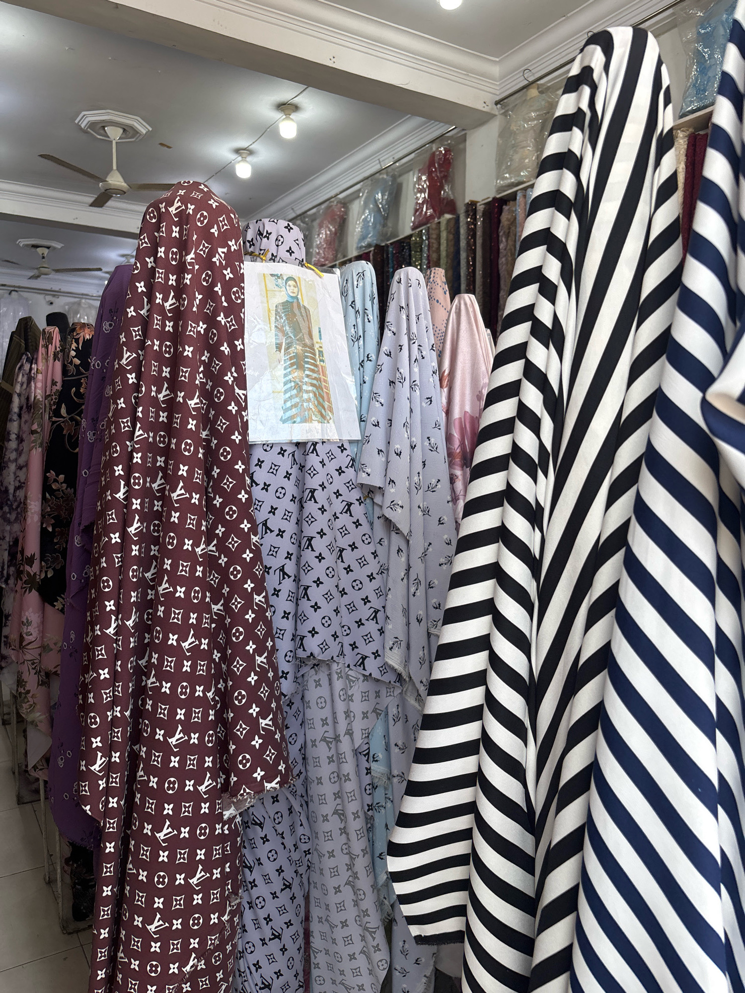 This image depicts the interior of a fabric store in Bireuen, Indonesia. The store is filled with various textiles displayed on racks and hanging from the ceiling. Prominently featured are several rolls of fabric, with the closest ones showcasing bold patterns: one with a repeating maroon Louis Vuitton-inspired monogram and another with black and white stripes. Behind these, other fabrics are visible, including light blue with a floral pattern and pastel pink. The environment appears to be a retail setting with fluorescent lighting and a ceiling fan, suggesting it is a typical shop. No people are clearly visible in the frame. The focus is on the diverse collection of fabrics available for sale.