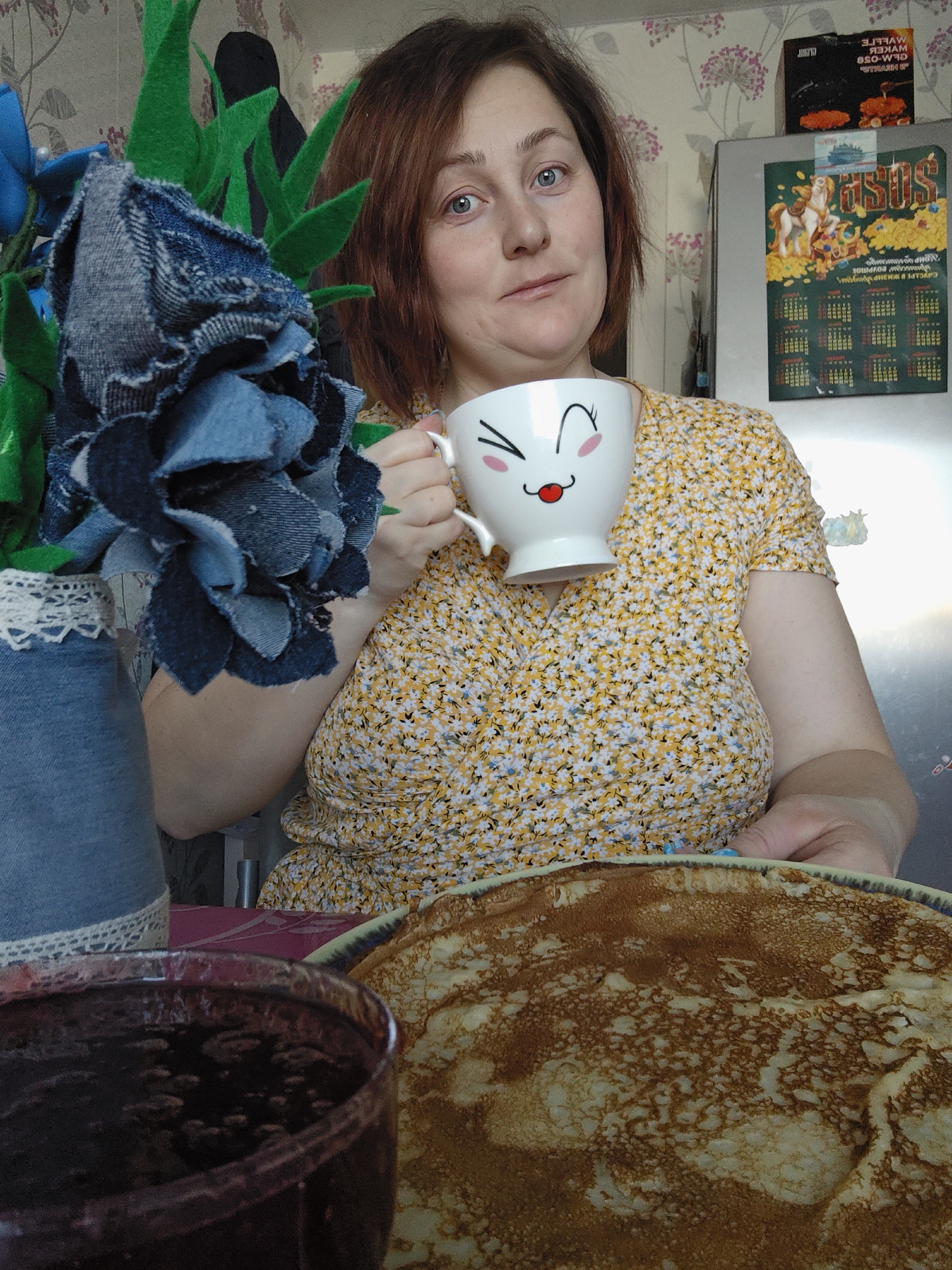 A woman with auburn hair and fair skin is holding a white mug with a cartoon face on it. She is wearing a yellow floral dress. In front of her is a plate of pancakes and a jar of dark jam. To the left of the woman is a vase made of denim with a denim flower in it. Behind the woman is a refrigerator with a calendar on it, which indicates the location of Markovskii, Russia. The room appears to be a kitchen.