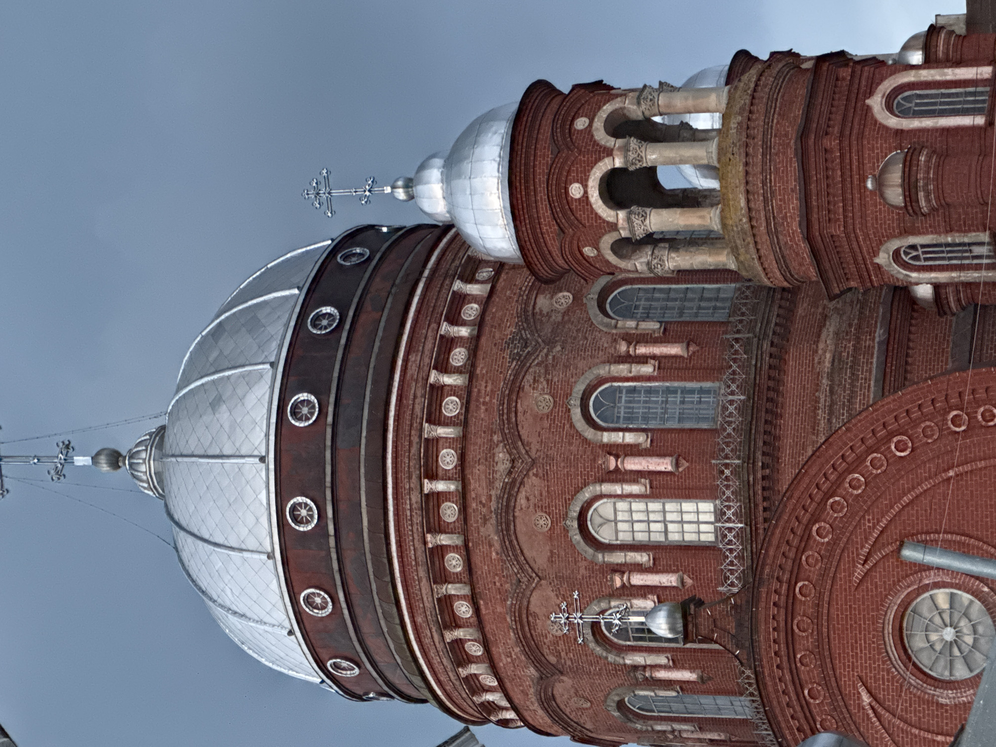 This is a close-up, low-angle shot of the domes and upper facade of the Ascension Cathedral, located in Urzhum, Russia. The cathedral is constructed primarily of red brick, with several silver domes. The largest dome, in the upper left of the frame, is ribbed and topped with a cross. Below it, a band of maroon separates it from the brickwork. A smaller, cylindrical tower with a silver dome and cross stands to the right of the main dome. This tower features an arcade of arches supported by columns. The brickwork of the cathedral is adorned with decorative elements, including rows of circular motifs and arched windows. The sky above is a pale, overcast blue. There are no people visible in the image.