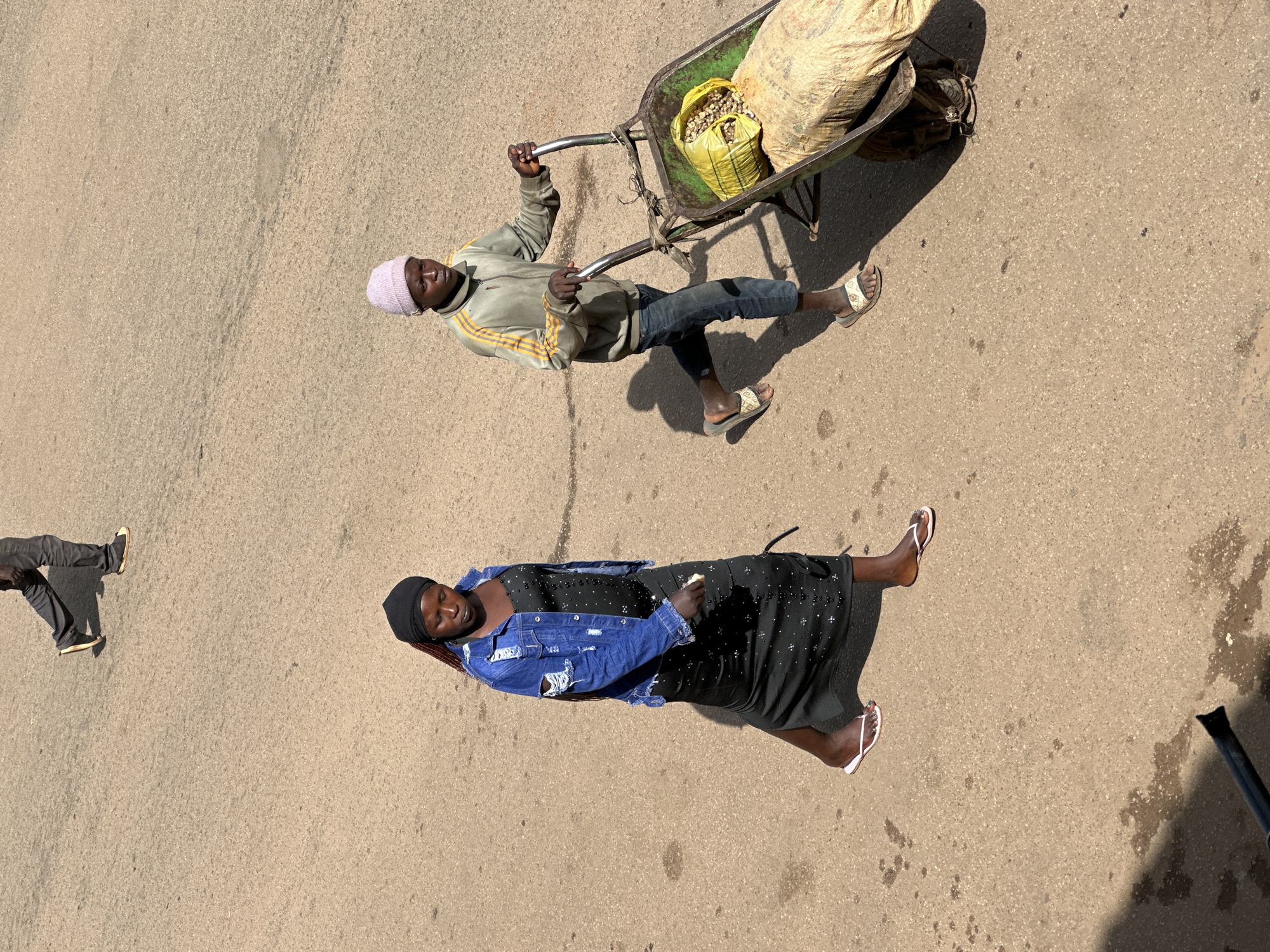 In Bukuru, Nigeria, two individuals are depicted on a paved surface. In the foreground, a young person wearing a light pink beanie, a grey and yellow jacket, and dark jeans is pushing a wheelbarrow. The wheelbarrow contains two large sacks, one of which is yellow and appears to be filled with a light-colored material, and another larger, tan sack. The person is wearing white and grey patterned sandals. To the left of the wheelbarrow pusher, a young woman is walking away from the viewer. She is wearing a black headscarf, a ripped denim jacket over a long black dress adorned with small white dots, and white flip-flops. Her hair, a braided dark color, is visible beneath her headscarf. In the upper portion of the image, the lower legs of another individual are visible, walking away from the viewer, wearing dark trousers and black shoes. The environment is a dry, dusty, paved area, likely a street or marketplace, with harsh sunlight casting shadows.