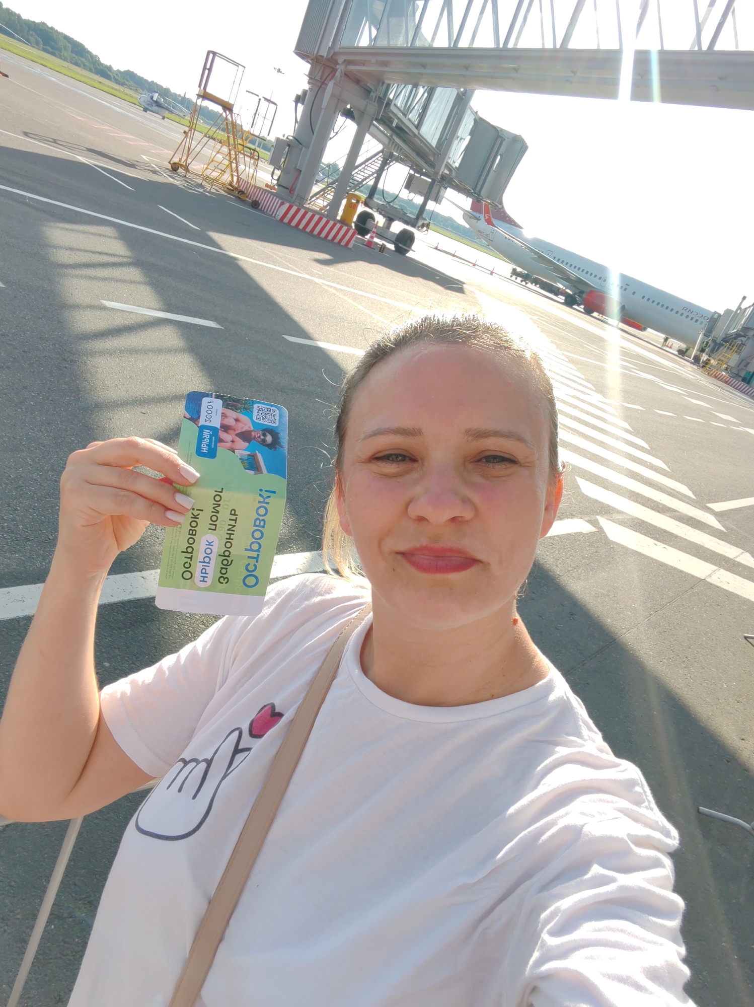 A woman is taking a selfie on an airport tarmac. She is wearing a white t-shirt with a heart gesture graphic on the sleeve and a beige crossbody bag. She is holding up a colorful ticket or boarding pass in her left hand. The ticket has Cyrillic text and images on it, including a picture of a man.
The background shows an airplane parked at an airport gate with a jet bridge extended to it. Other airport infrastructure, such as stairs and barriers, are also visible. The scene is outdoors on a sunny day, indicated by the bright light and long shadows. The time of day appears to be late afternoon or early evening due to the angle of the sun. The overall atmosphere is cheerful and suggests the woman is about to embark on a trip. The location context provided is Khrabrovo, Russia, which is likely the airport where this photo was taken.
The text on the ticket is partially legible. It includes "ОСЛІВОВОКІ", "HPÏbok", "UOWOL", and "завдонцр" which appears to be Ukrainian.
