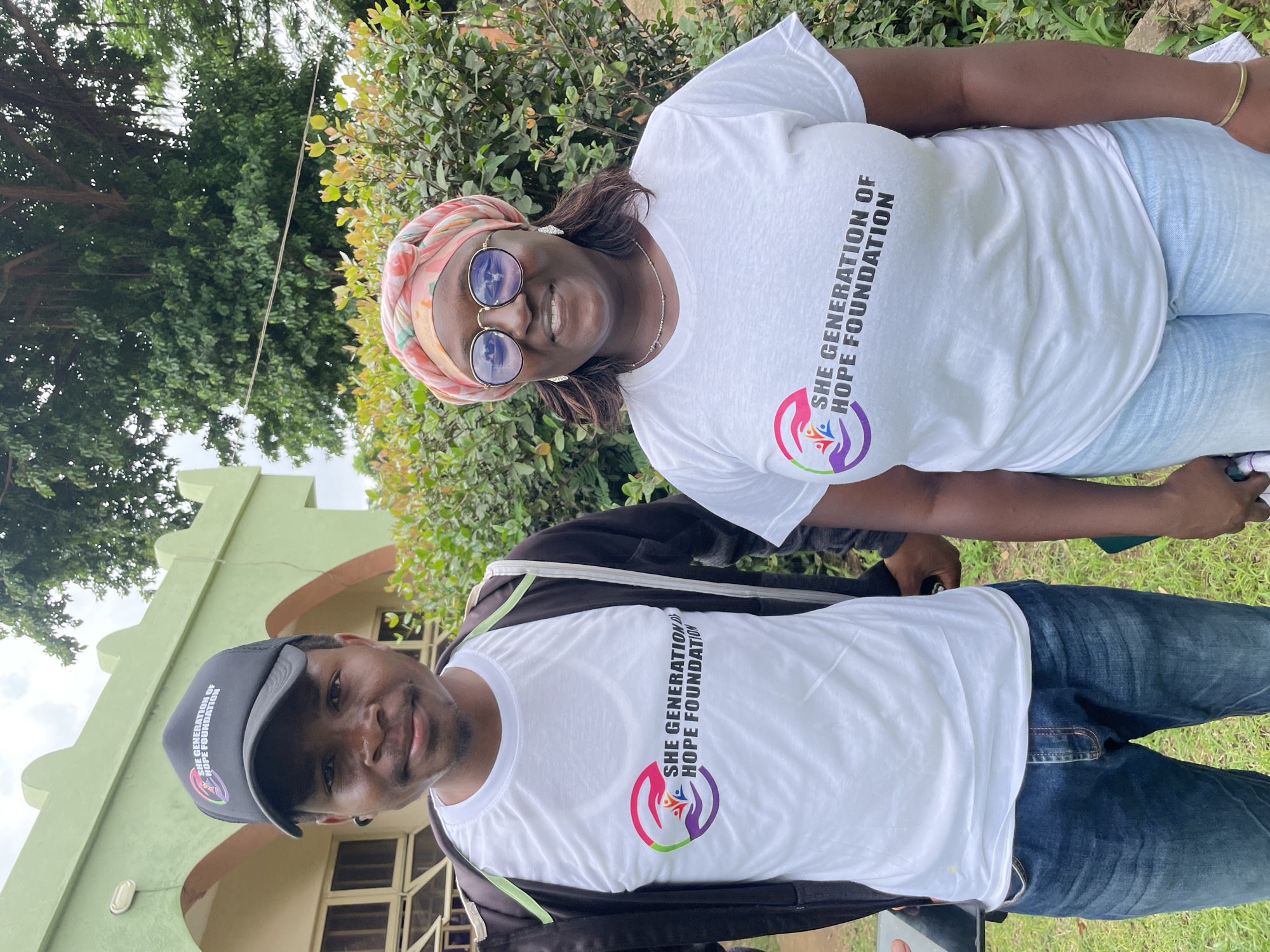 Two individuals, a man and a woman, stand outdoors in front of a light green building with arched doorways and windows. The man, on the left, wears a black baseball cap with a logo, a white t-shirt with the "SHE GENERATION OF HOPE FOUNDATION" logo, and a black zipped jacket over it. He is also wearing blue jeans. The woman, on the right, is wearing a headscarf, round glasses, earrings, a necklace, a white t-shirt with the same "SHE GENERATION OF HOPE FOUNDATION" logo, and light blue jeans. They are both smiling at the camera. Lush green foliage and a large tree with hanging roots are visible behind them. The ground is covered in grass. The building appears to be in a residential or institutional area within Unguwar Shanu, Nigeria.