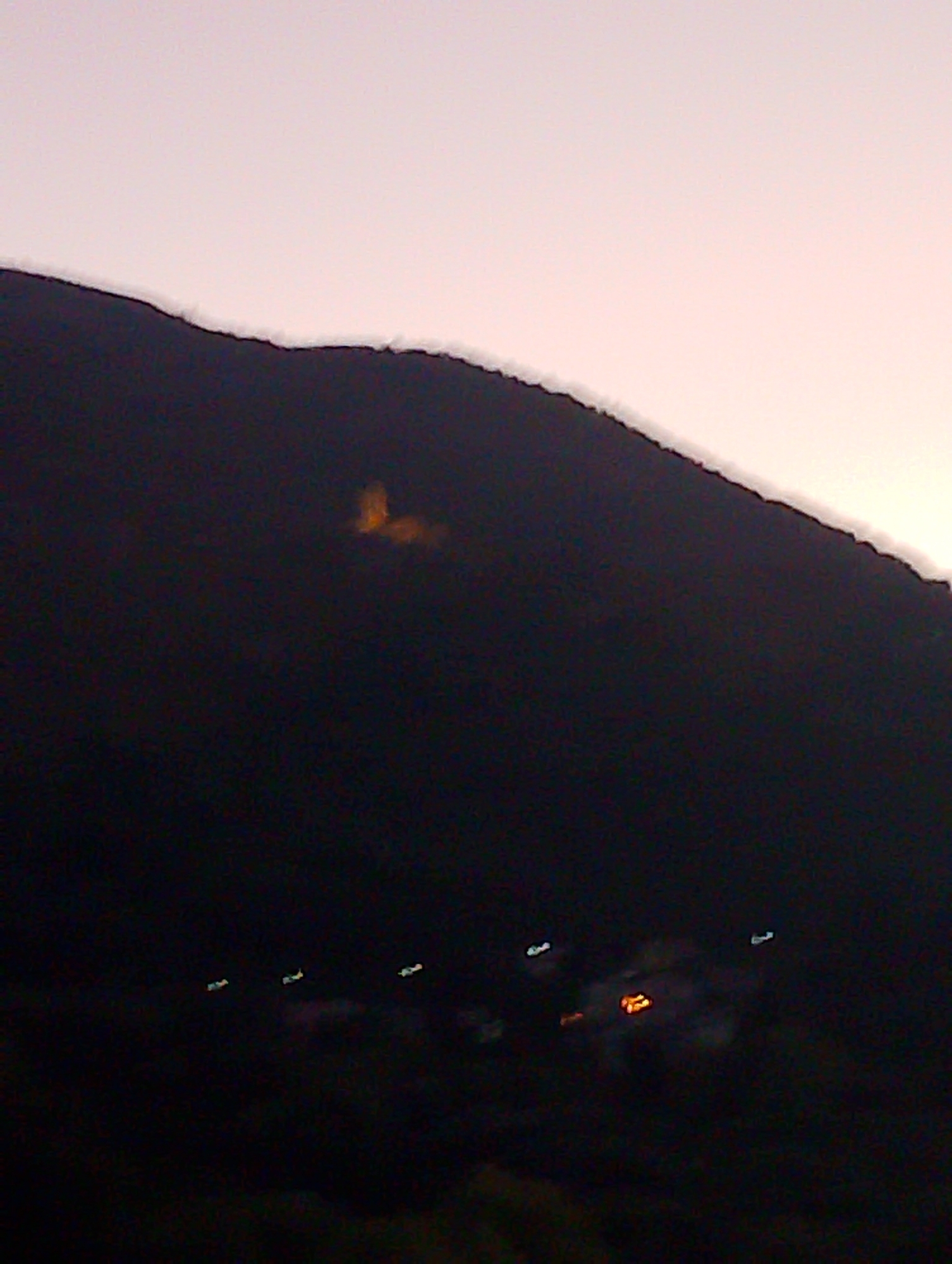 The image shows a dark, mountainous landscape with a pale sky at dusk or dawn. A prominent feature is a cluster of warm, orange lights on the side of the mountain, which could be a small settlement or a fire. Below this, near the base of the mountain, are several faint, elongated lights that appear to be from vehicles on a road. The overall impression is one of a rural or semi-rural setting at night, with human activity indicated by the lights. The specific location in Fiamignano, Italy, is not explicitly visible but the context suggests it is a view of the surrounding mountainous terrain.