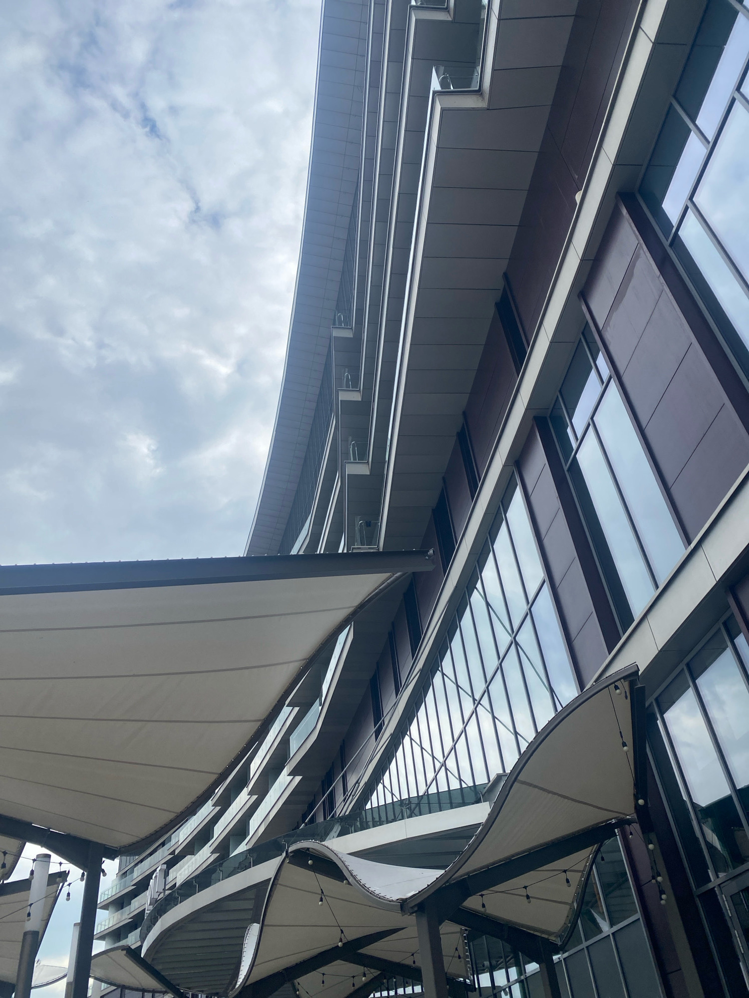 The image shows the facade of a modern building, likely a hotel or residential complex, in Minsk, Belarus. The building has a contemporary design with large glass windows and dark brown paneling. The perspective is from below, looking up at the structure against a cloudy sky. In the foreground, a series of white fabric awnings are visible, supported by dark metal poles. String lights are hung under these awnings, suggesting an outdoor seating or event area. The building itself features multiple balconies with glass railings, indicating its upper floors are used for accommodation or offices. The overall impression is one of urban architecture and design.