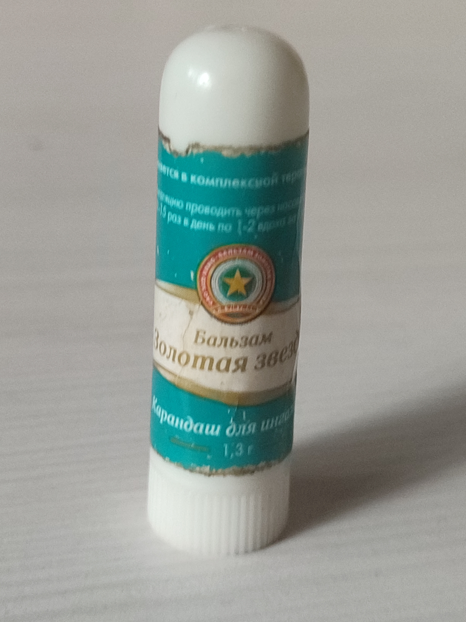 A white, bullet-shaped lip balm stick with a teal label is centered on a light-colored, textured surface. The label features text in Russian and a circular emblem with a star. The text is partially obscured but includes the words "Бальзам" (Balsam), "Золотая Звезда" (Golden Star), and "Карандаш для" (Pencil for). The bottom of the stick has a ribbed white cap. The background is a plain, soft grey. The image does not contain any information related to the city of Kungur, Russia.