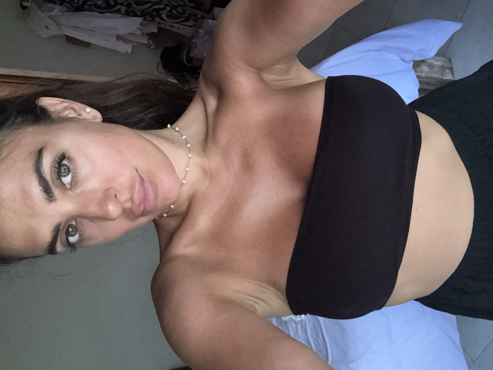 A woman is taking a selfie indoors. She is wearing a black strapless top and a pearl necklace. She is looking directly at the camera. A blue pillow is visible behind her. Clothes are visible in the background. The location is not discernible from the image. The city of North Kuta, Indonesia, cannot be confirmed.