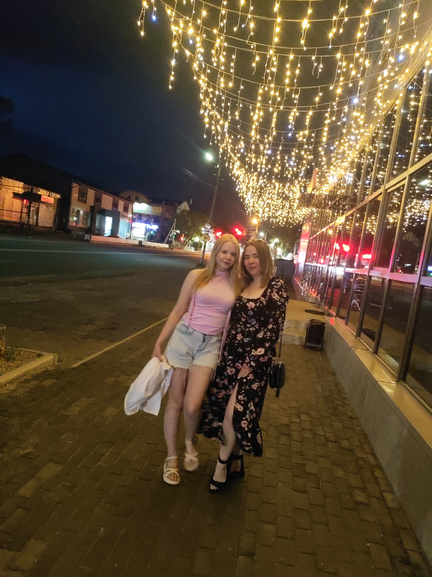 Two women stand on a brick-paved sidewalk at night. The woman on the left has blonde hair, a light pink tank top, and light denim shorts. She is holding a white garment. The woman on the right has brown hair and wears a black dress with a floral pattern and a high slit. She carries a black purse over her shoulder. They are positioned under a canopy of string lights that illuminate the scene. Behind them, across a street, are several buildings with illuminated windows and some signage, suggesting an urban commercial area. The buildings appear to be two or three stories high, with one having a facade of light-colored brick and another with darker elements. Streetlights and traffic lights are visible in the background, indicating an active urban environment. The overall atmosphere is festive due to the abundant lighting. This scene is located in the city of Shimanovsk, Russia.