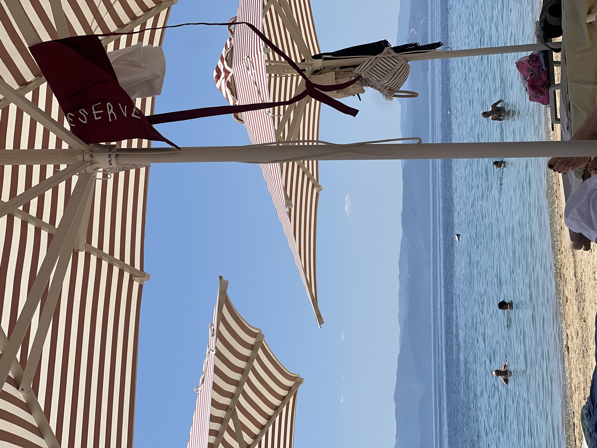 This image captures a sun-drenched beach scene in Troulos, Greece, with clear blue skies and calm, azure waters. In the foreground, striped beach umbrellas, predominantly in shades of brown and white, provide shade. One umbrella features a maroon accessory with white lettering that reads "RESERVE," and several garments, including a bikini top and what appears to be a white towel, are casually hung from its pole. Another umbrella is visible in the middle ground, also striped, and a third is angled in the upper left of the frame.
The sea is populated by several individuals. Two men are visible in the water, one closer to the shore and another further out. A woman is also in the water, and another person's head and shoulders are just above the surface. A yellow buoy is floating in the distance. The shoreline is sandy, and lounge chairs with towels are visible on the beach. In the far distance, a range of hills or mountains forms the horizon. The overall impression is one of a relaxed and idyllic beach day.