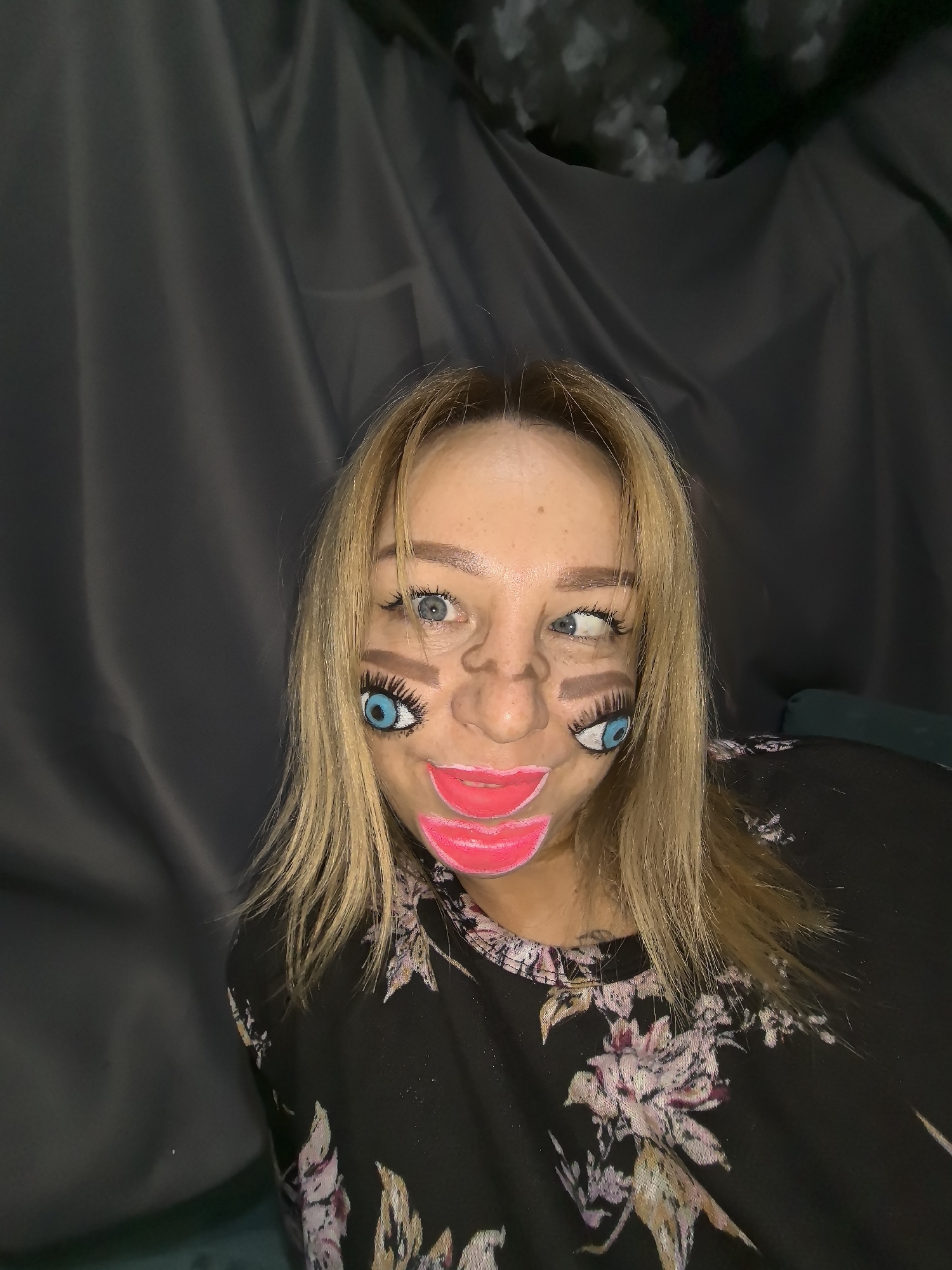An indoor portrait features a woman with blonde hair and blue eyes, showcasing an elaborate and whimsical face paint design. Her actual face is adorned with exaggerated, cartoon-like features. Two large, bright blue eyes with prominent black eyelashes are painted on her cheeks, positioned below her natural eyes, which are visible and looking to the side. A stylized, slightly comical nose is painted over her real nose. Below this, a large, glossy pink mouth with distinct lips is painted, extending from her upper lip down towards her chin. Thick, brown painted eyebrows are visible above the drawn eyes.
The woman is wearing a dark, long-sleeved top with a vibrant floral pattern featuring shades of pink, white, and hints of brown. Her blonde hair is parted on the side and falls around her shoulders.
The background consists of dark grey or black draped fabric, possibly a curtain or blanket, creating a somewhat uniform setting. In the upper right corner, there are indistinct lighter shapes, possibly blurred objects or more fabric. A glimpse of a dark green or teal upholstered surface, likely a cushion or furniture, is visible on the left side of the frame, suggesting she is seated.
The overall impression is one of artistic expression, playfulness, or preparation for an event like a costume party. The lighting appears to be artificial, indicating an indoor setting, possibly at night. No visible text is present in the image. The location context provided is Noviy, Russia, though no specific visual cues in the image confirm this.
